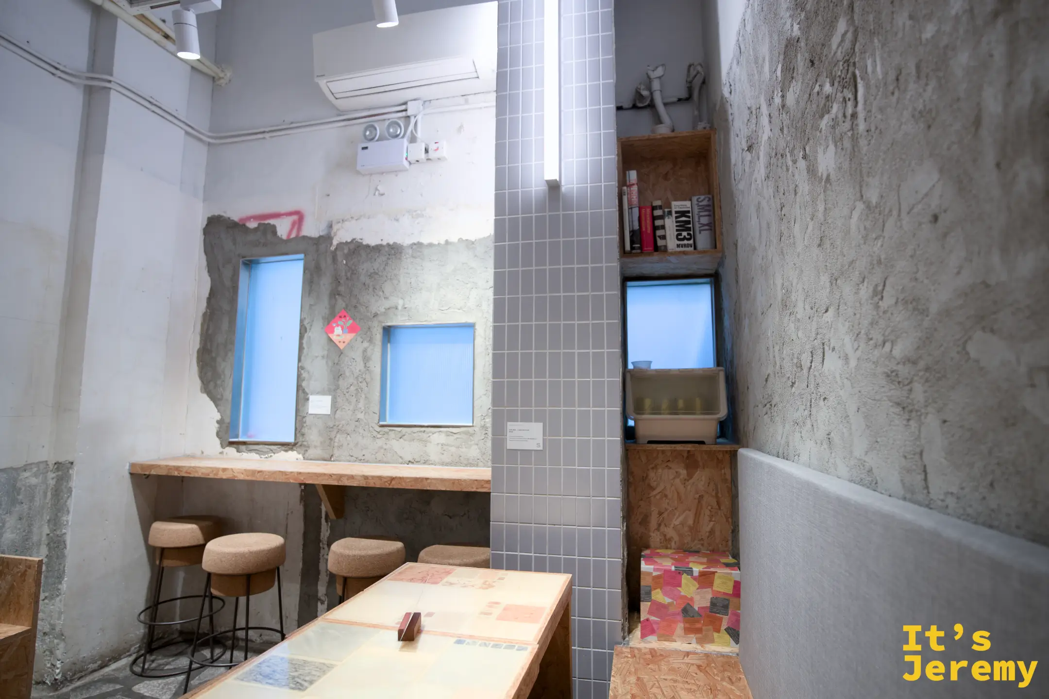店內工業風空間，裸露水泥牆保留斑駁質感，牆上開著藍色小窗，靠牆吧台配軟木塞圓凳，中央一張木質長桌
