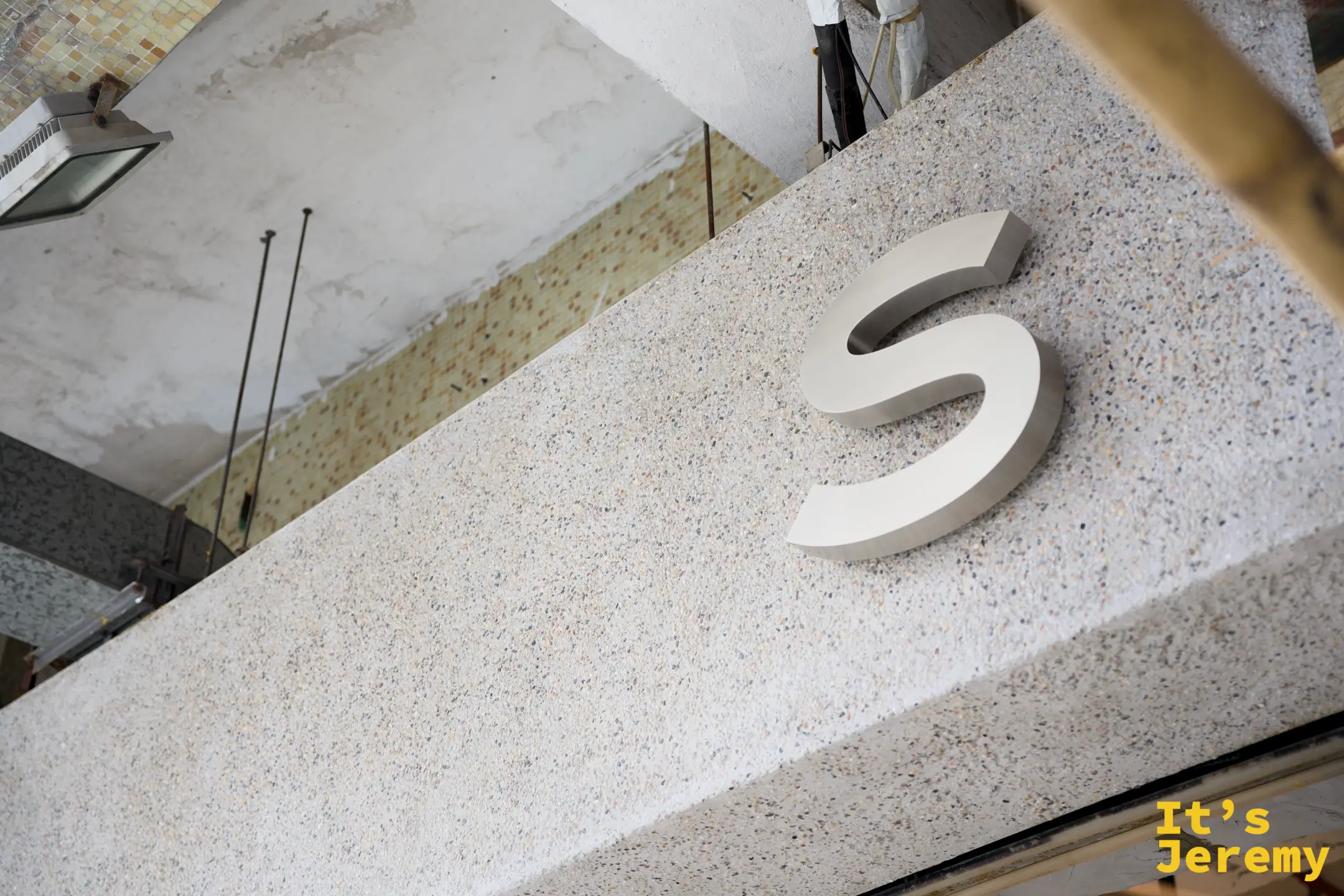 仰角拍攝石砌外牆上一顆立體銀色的「S」字母招牌，牆面是水磨石質感