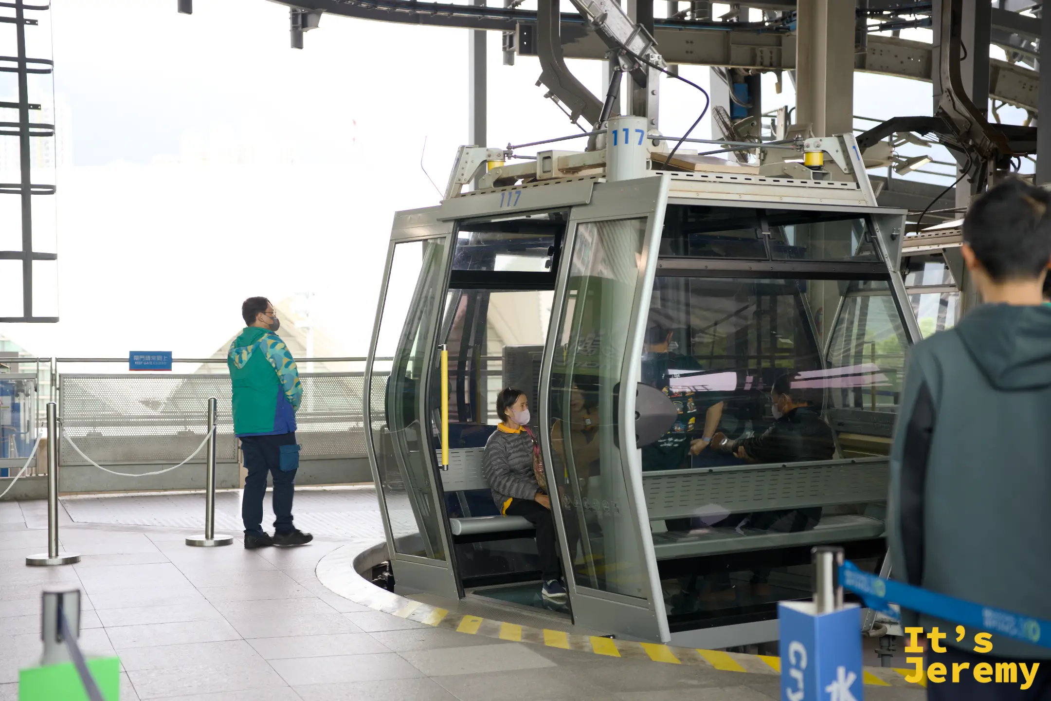 昂坪 360 月台邊，一名穿藍綠色制服的站務人員站在車廂旁，銀色纜車車廂門敞開，車內坐著戴口罩的乘客