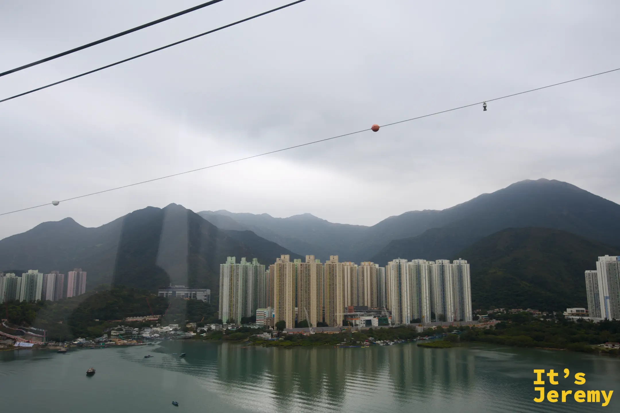 從纜車車廂內往外看，碧綠的東涌灣對岸是一整排高聳的住宅大樓，背後是纏著薄霧的大嶼山群山，纜索上遠遠掛著一台紅色車廂