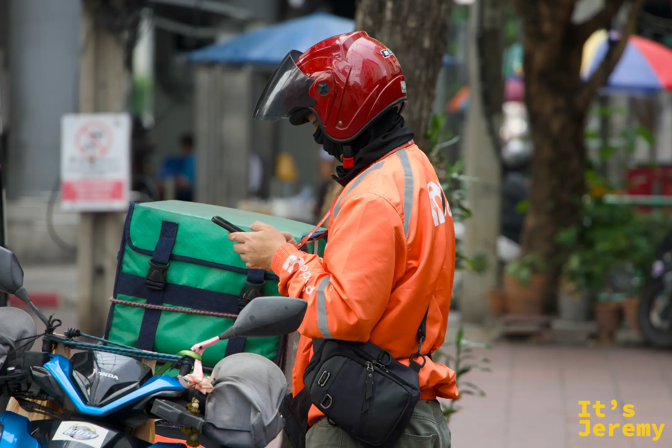 橘色制服的 Shopee Food 外送員站在路邊低頭滑手機，吞武里的街頭節奏跟 Siam 不是同一個時區