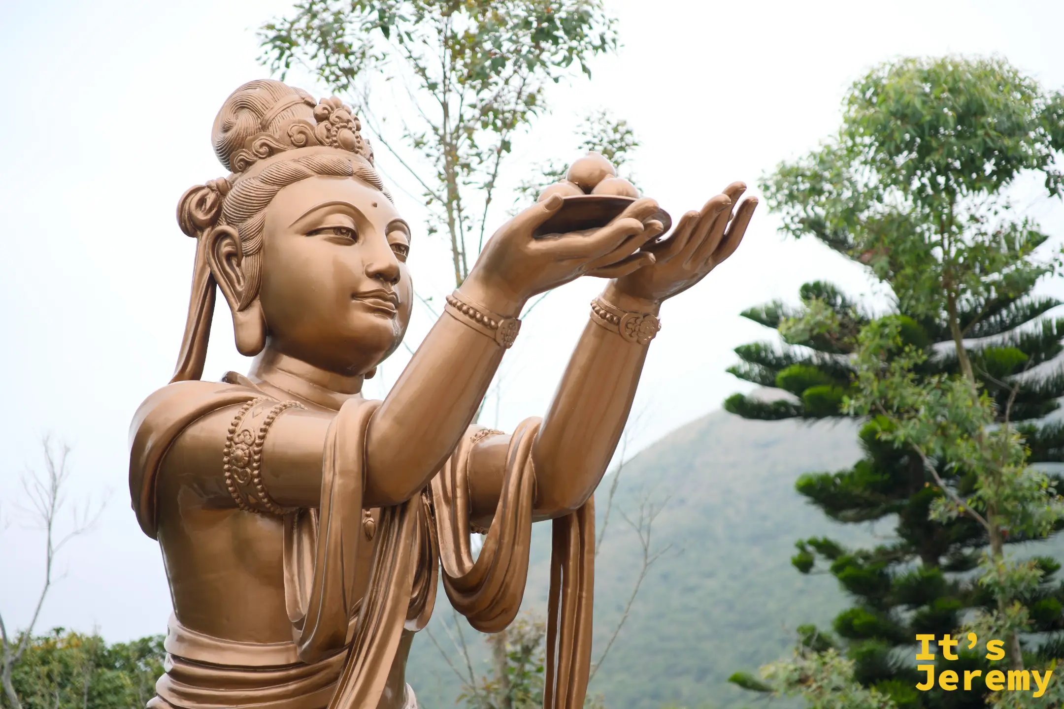 青銅天母像雙手捧起供果向前獻上，衣飾線條流暢，背景是遠處的大嶼山群山
