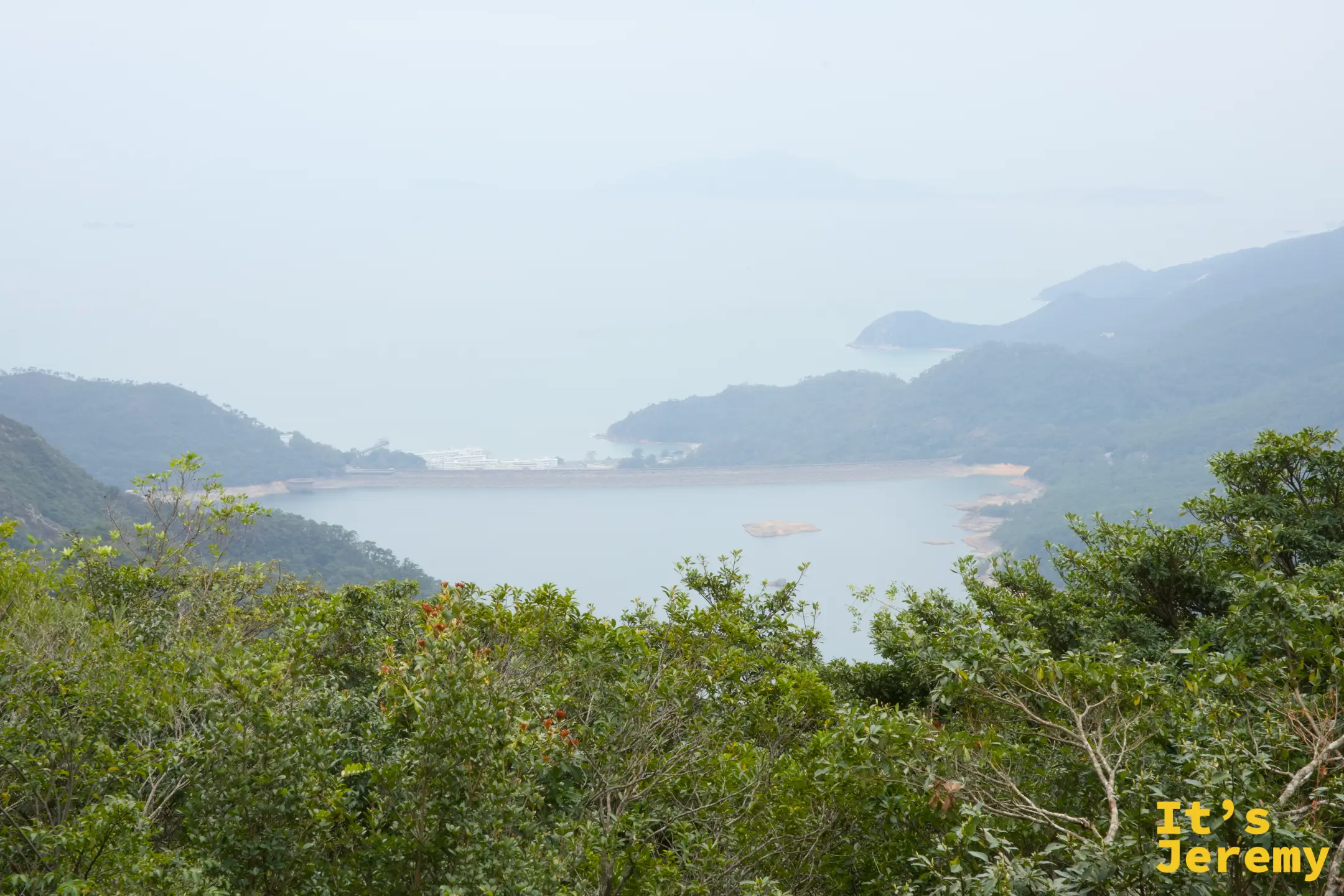 從心經簡林步道高處眺望，山谷裡一泓碧綠水塘，後方是層層山巒與霧氣氤氳的海灣離島，前景是茂密綠色灌木