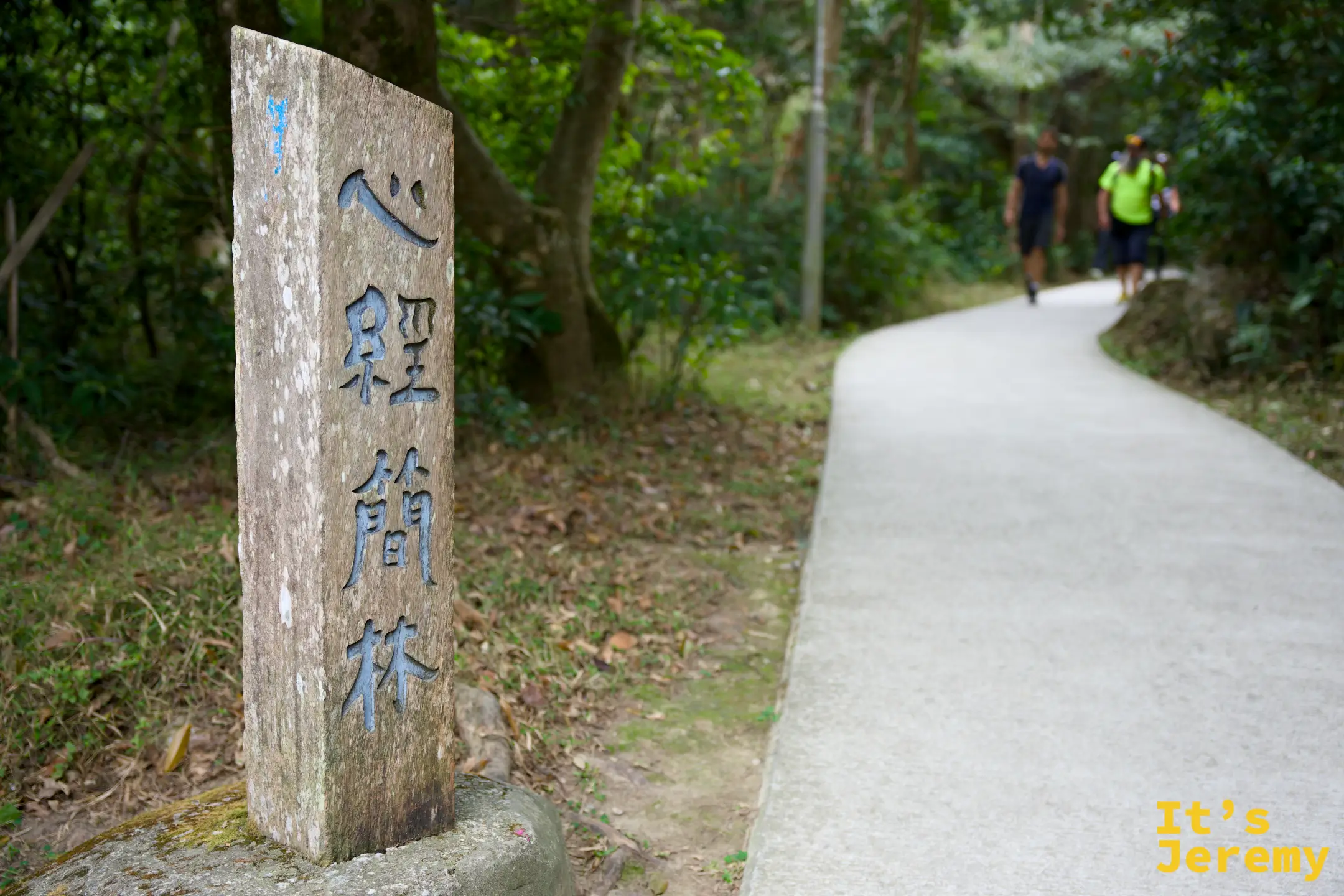 路邊立著刻有「心經簡林」四字的石碑，右側是一條平緩的水泥步道，兩名健行者背著背包往前走，四周綠樹環繞