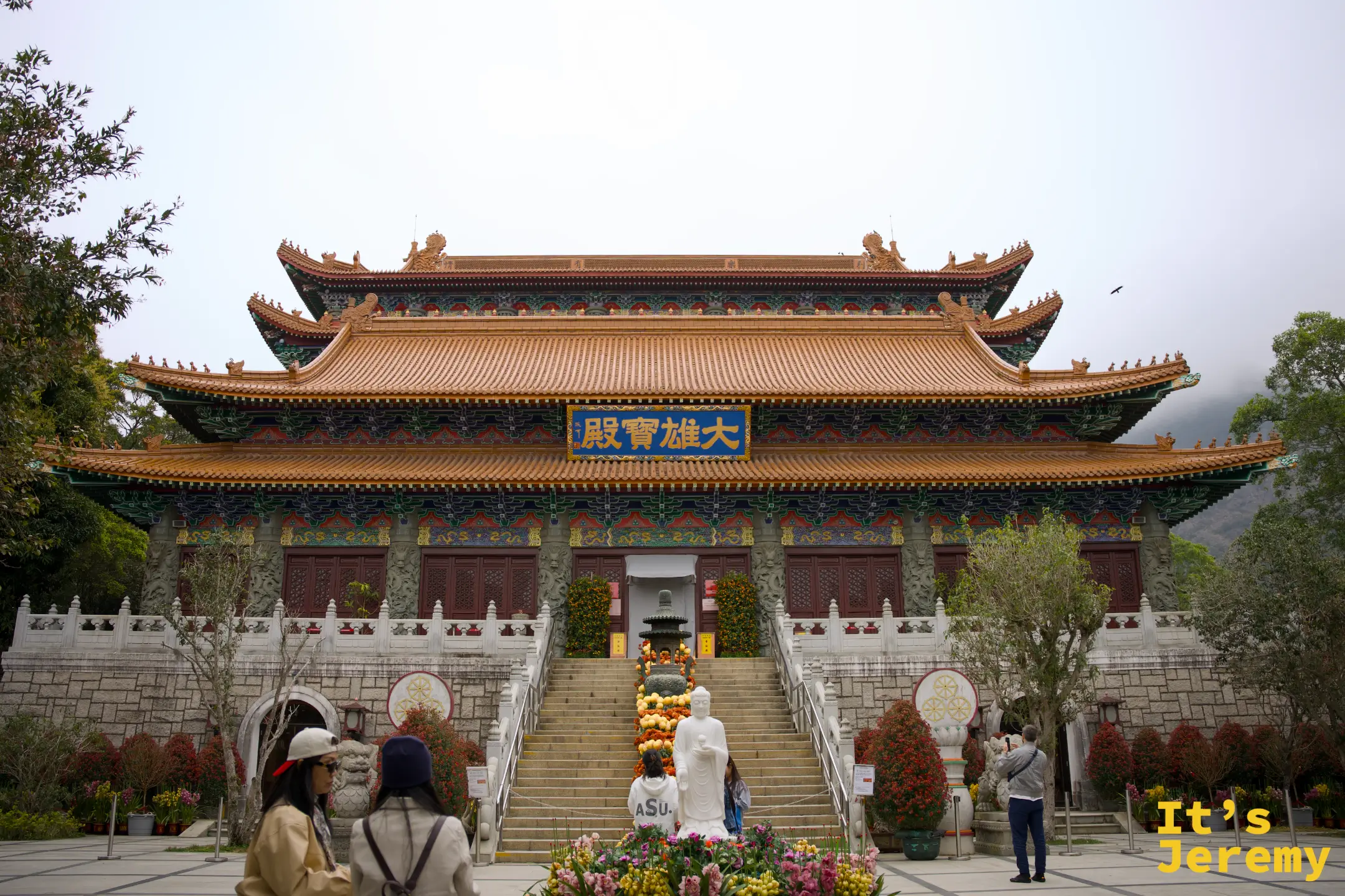 寶蓮禪寺大雄寶殿正面，橙紅琉璃瓦頂、藍底金字匾額，殿前白石階梯兩側擺滿菊花盆栽，遊客在前方參觀