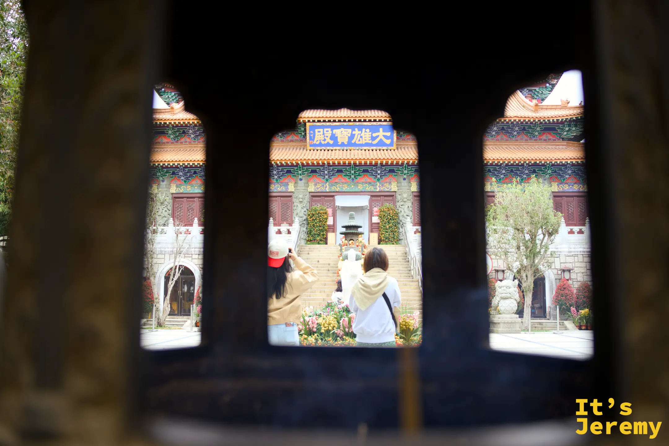 透過巨大銅香爐的鏤空窗格望向大雄寶殿，藍底金字「大雄寶殿」匾額被框在窗格中，前方有兩名遊客在殿前拍照