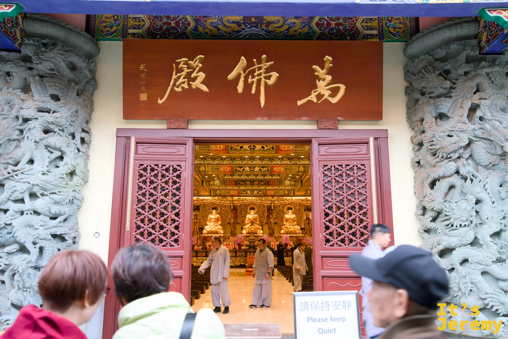 萬佛殿入口，紅底金字「萬佛殿」匾額高掛，兩側石柱雕著盤龍，門內可見金色佛壇與佛像，門口有僧人與遊客