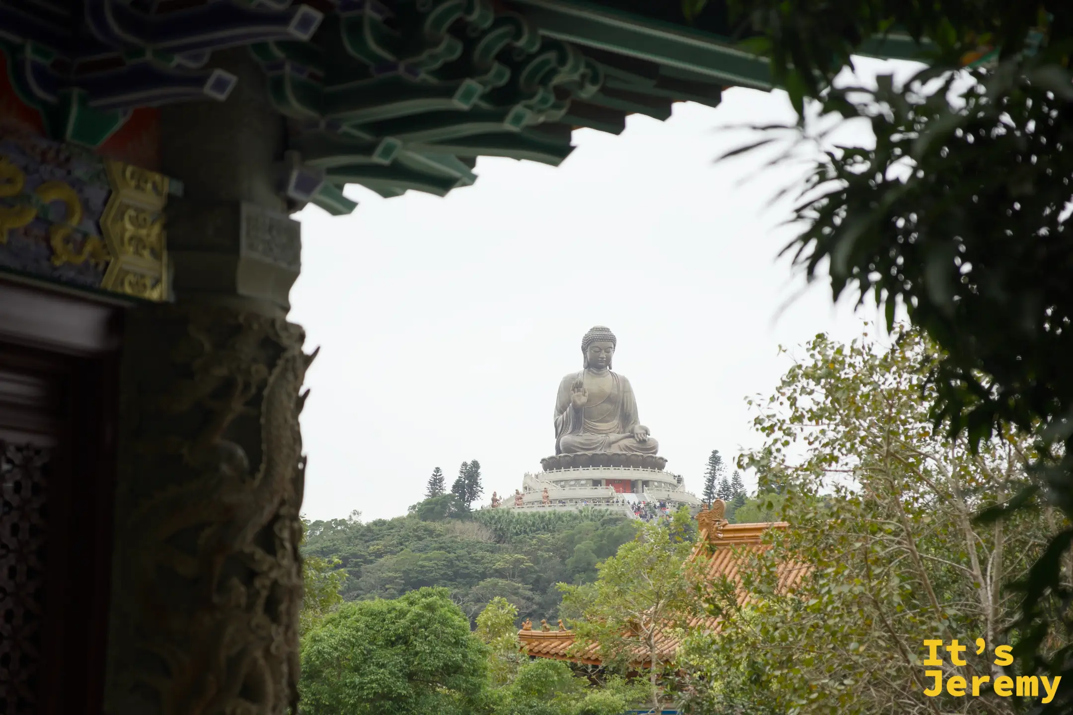 從寶蓮禪寺殿堂的彩繪飛簷下望出去，遠處山頭上天壇大佛端坐在綠樹之上，飛簷與大佛同框