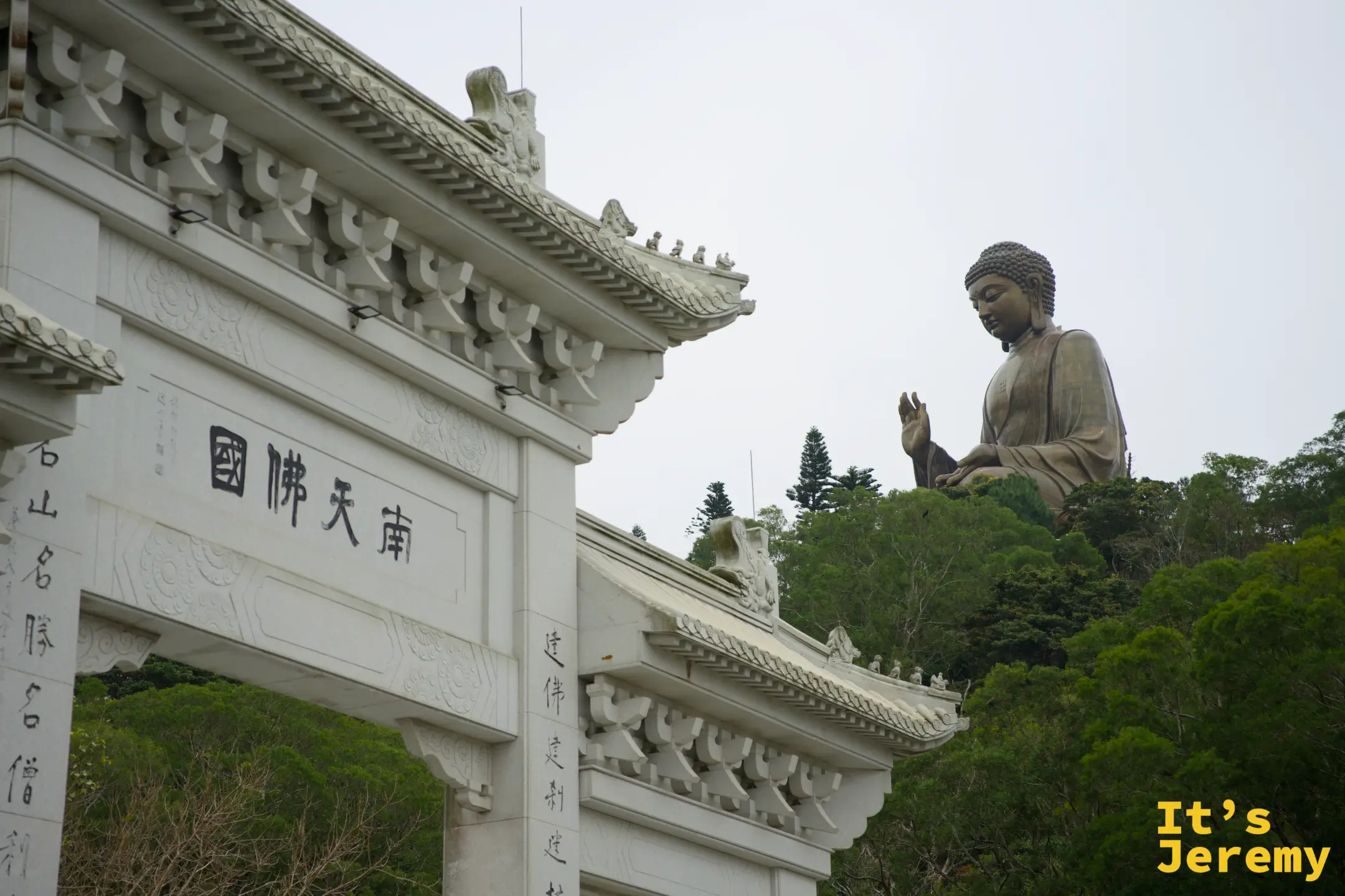 白色石造「南天佛國」牌坊立在前景，牌坊右後方的山頭上，天壇大佛端坐在綠樹之上，天色灰白