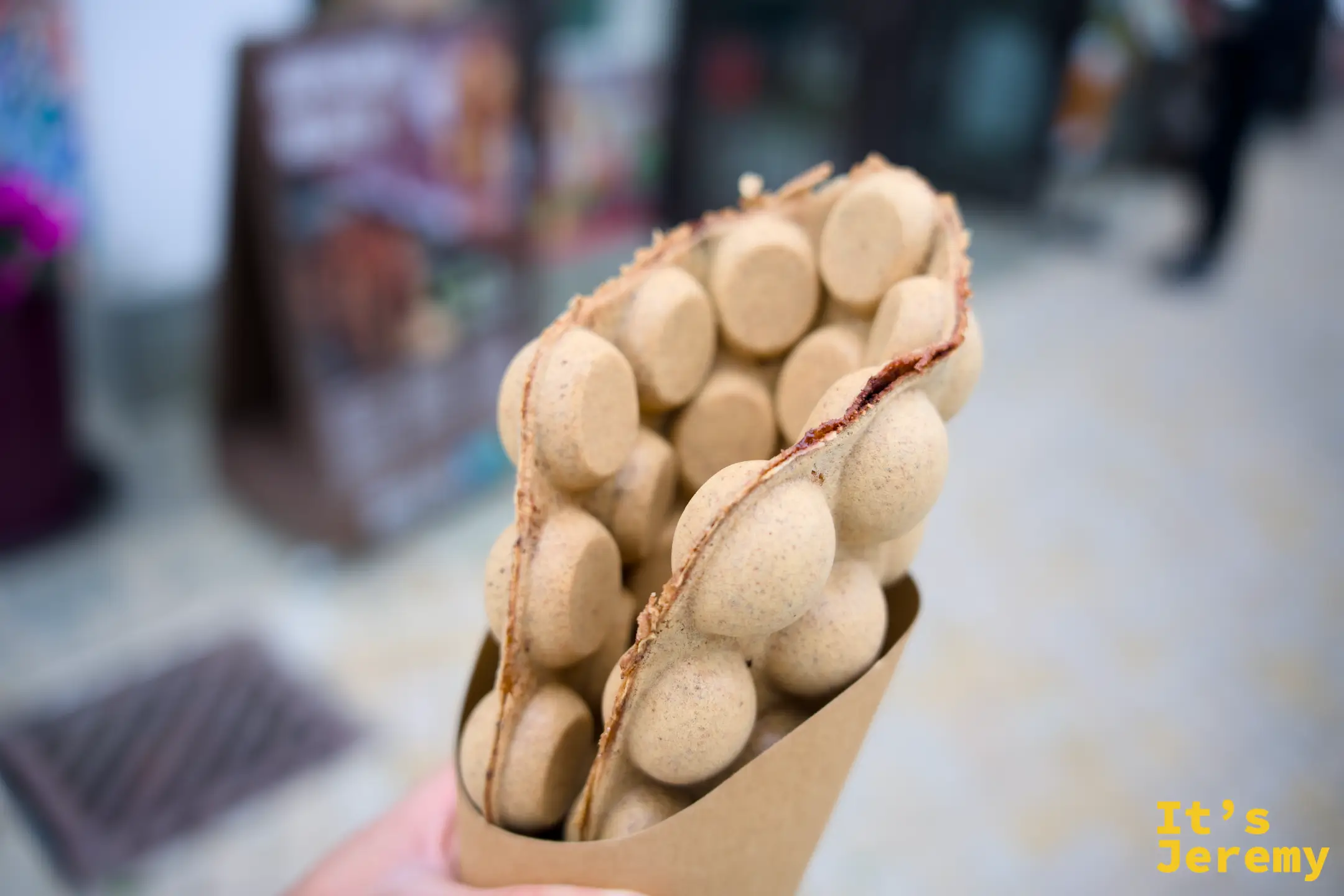 手拿一份牛皮紙包著的港式雞蛋仔，一顆顆鼓圓的蛋泡烤成淡金色，邊緣帶著焦香，背景是昂坪市集的店面
