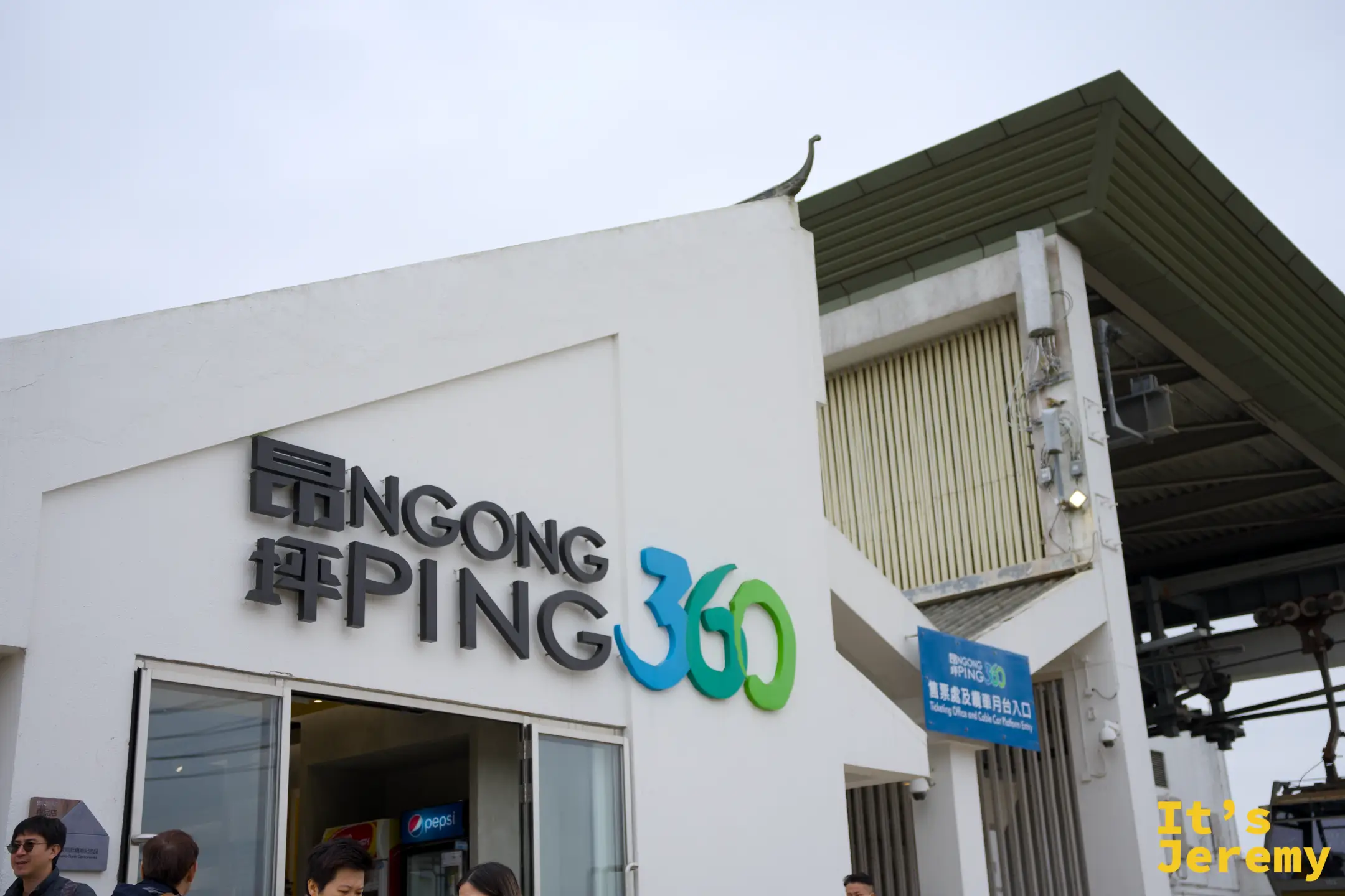 昂坪 360 纜車站白色站體外觀，牆上有藍綠色「昂坪 PING 360」字樣，站外聚集許多遊客，纜車車廂正沿纜索滑進站