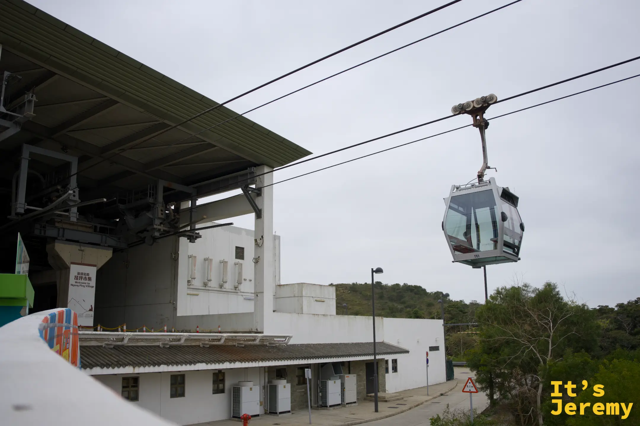 仰角拍纜車車廂沿纜索滑進站，車廂懸掛在纜索上，下方是白色站體與綠色遮陽棚，遠處是大嶼山山丘