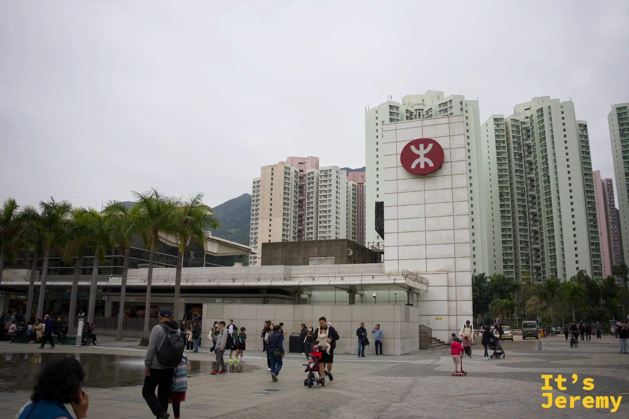 東涌港鐵站白色站體外觀，牆上是紅色港鐵標誌，站前廣場開闊，人來人往，背後是連綿青山與住宅大樓