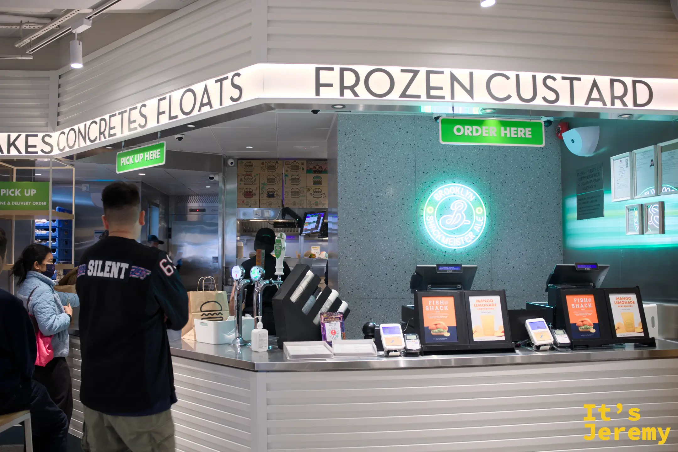 SHAKE SHACK 店內點餐櫃檯，上方掛著 SHAKES CONCRETES FLOATS 與 FROZEN CUSTARD 燈牌，牆上有綠色霓虹漢堡 logo，一名顧客站在櫃檯前等候