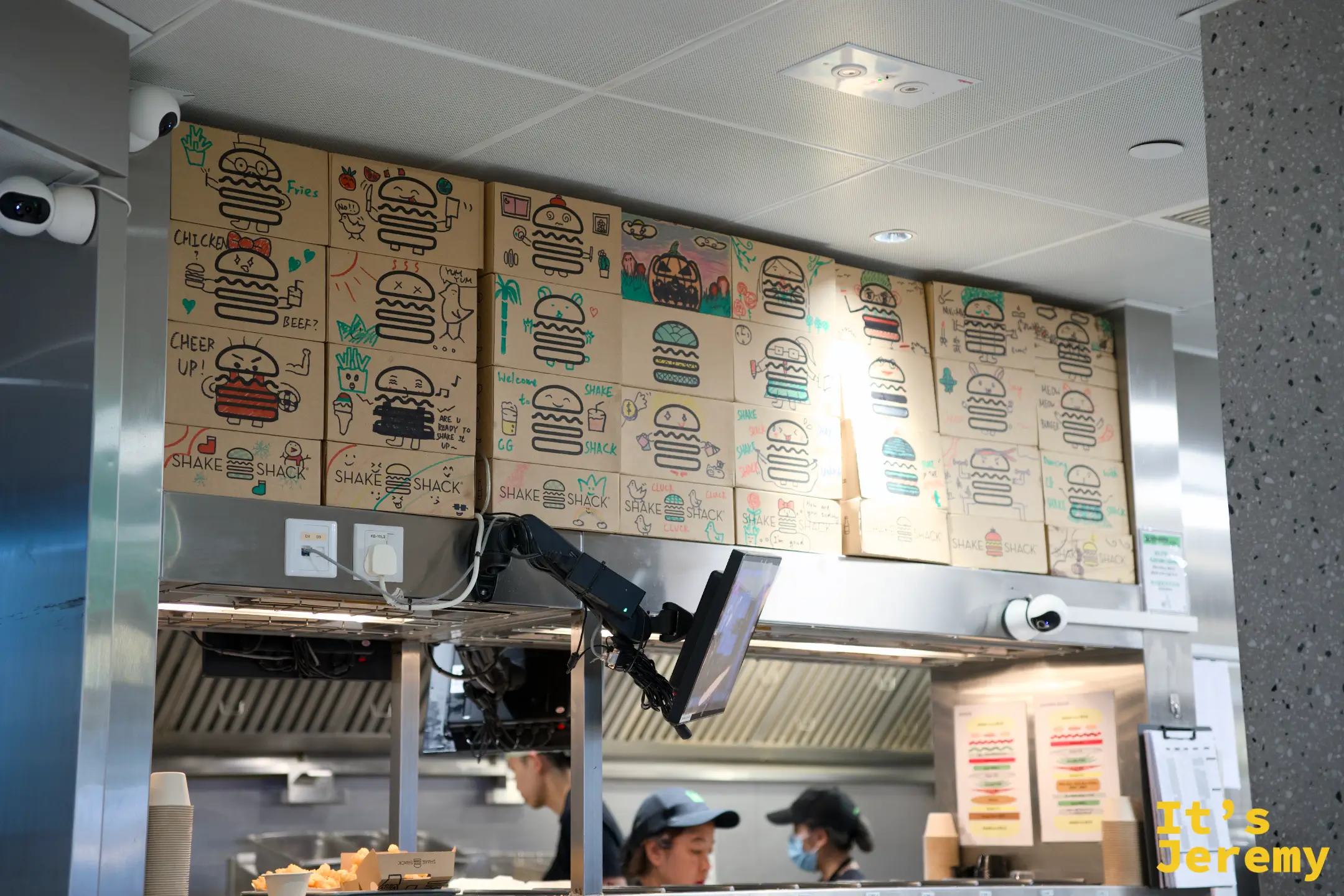 SHAKE SHACK 廚房上方堆疊著一整面牆的牛皮紙外帶紙盒，每個盒子都被手繪上不同的漢堡塗鴉與表情，廚房內店員正在備餐
