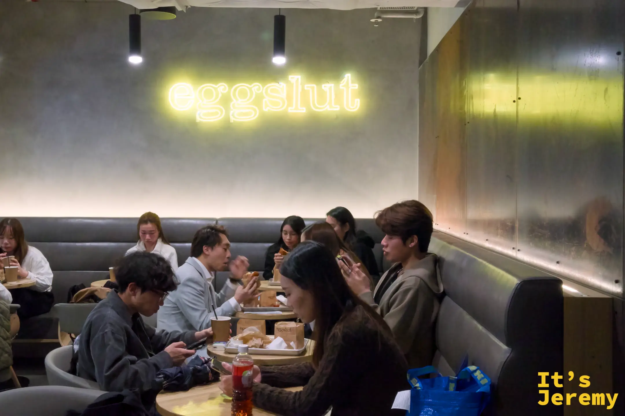 eggslut 店內用餐區，水泥牆上掛著黃色「eggslut」霓虹燈，皮革卡座坐滿客人，桌上放著食物與飲料