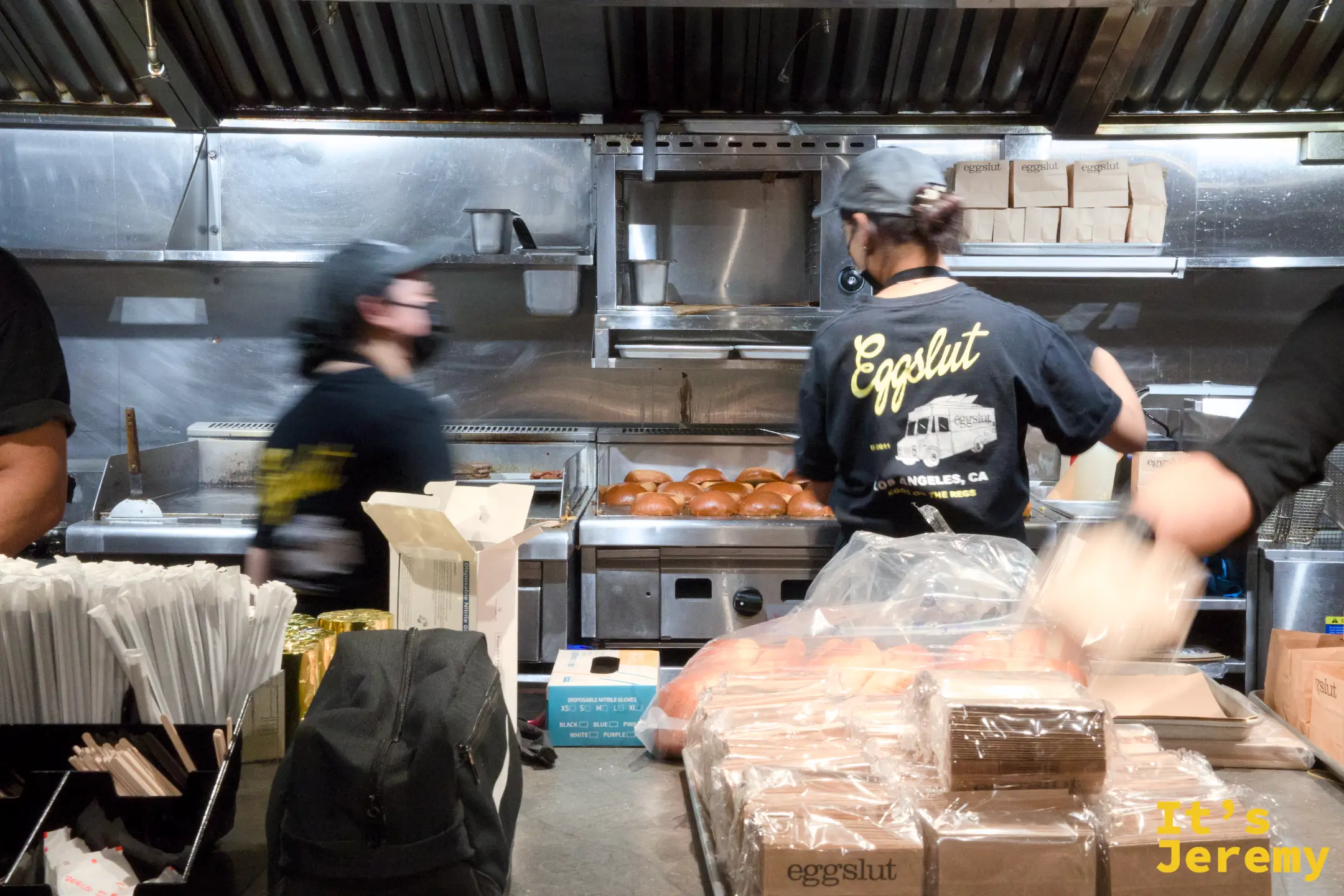 eggslut 半開放式廚房，店員穿著印有洛杉磯餐車圖案的黑色制服在爐台前備餐，檯面上排著一列布里歐麵包與外帶紙袋