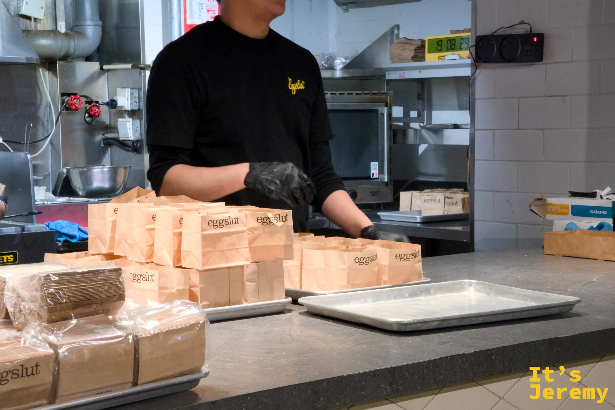 eggslut 出餐檯前，一名戴手套的店員正把三明治裝進印有 eggslut 字樣的牛皮紙袋，檯面上排著一列紙袋