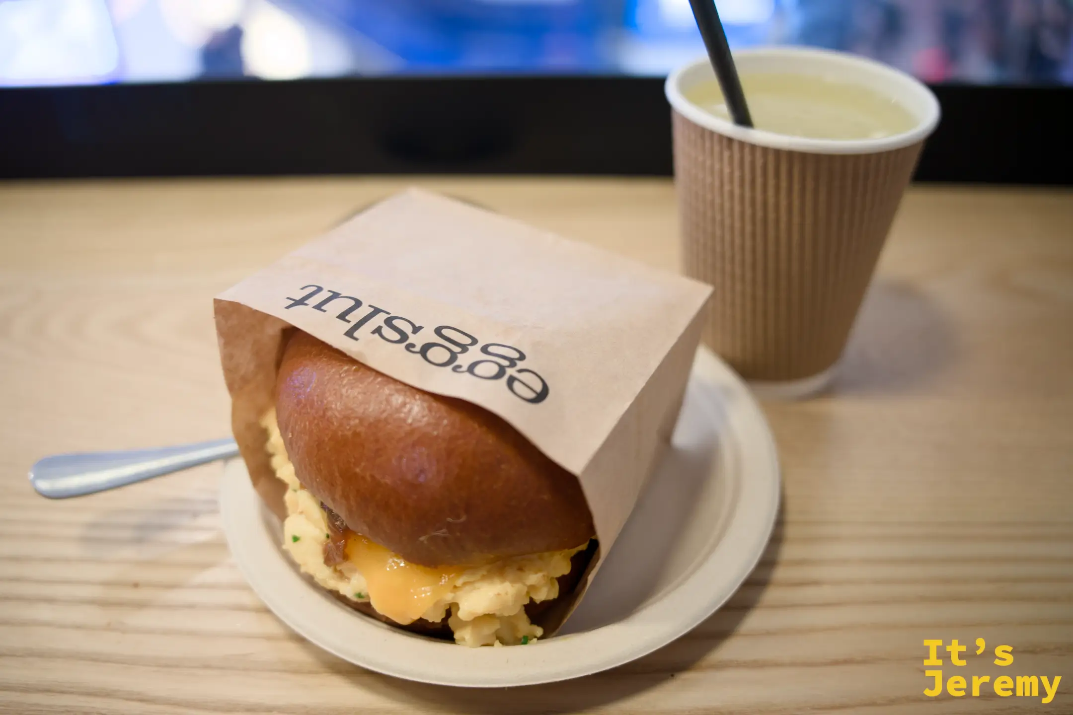 eggslut 牛皮紙套包著的 Fairfax 炒蛋三明治放在白盤上，露出滑嫩的炒蛋與焦糖洋蔥，旁邊放著一杯插著吸管的飲料