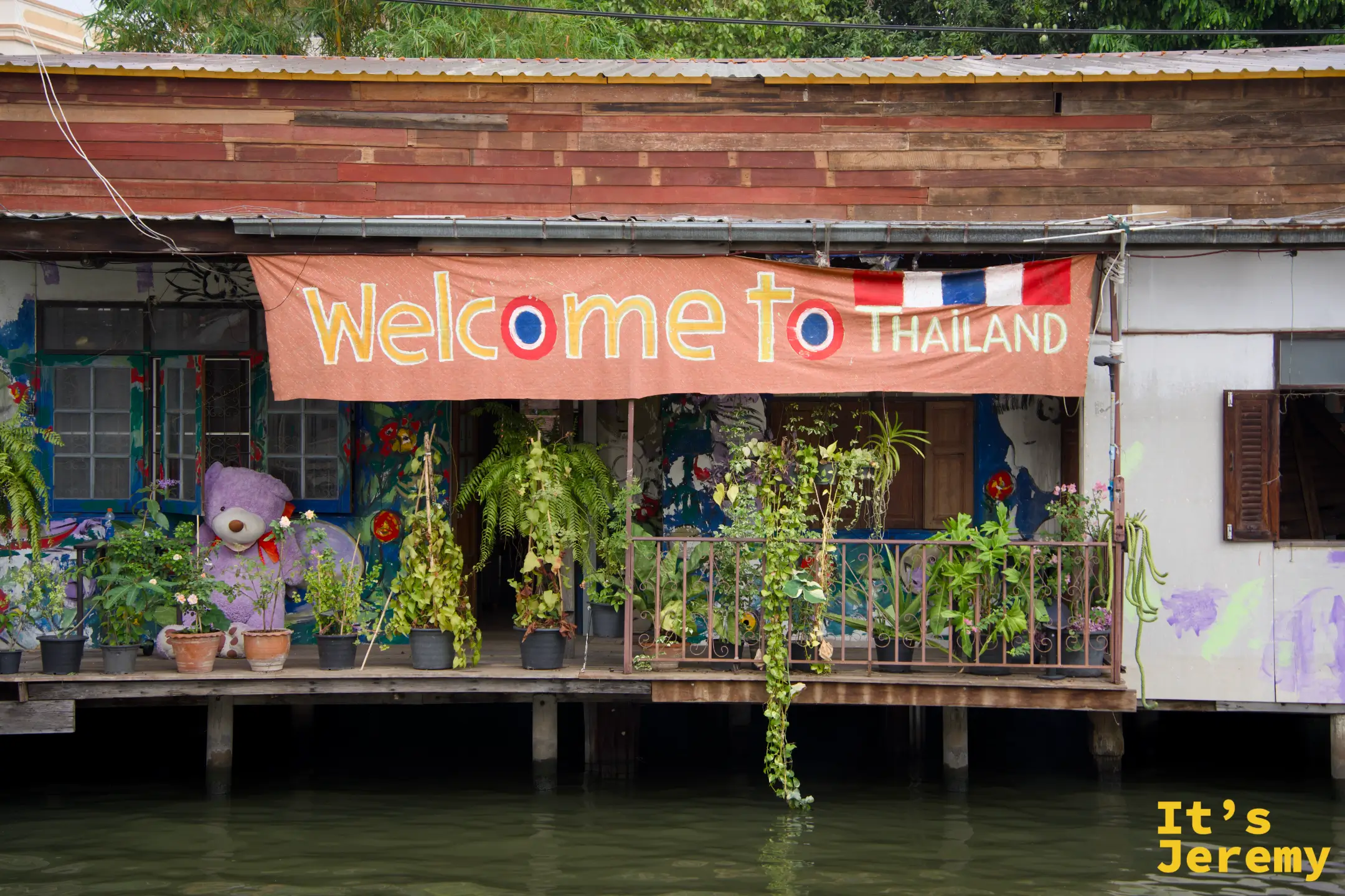 從長尾船上回望運河旁的木造老宅，掛著「Welcome to Thailand」布條，這一段是整趟最有感的視角
