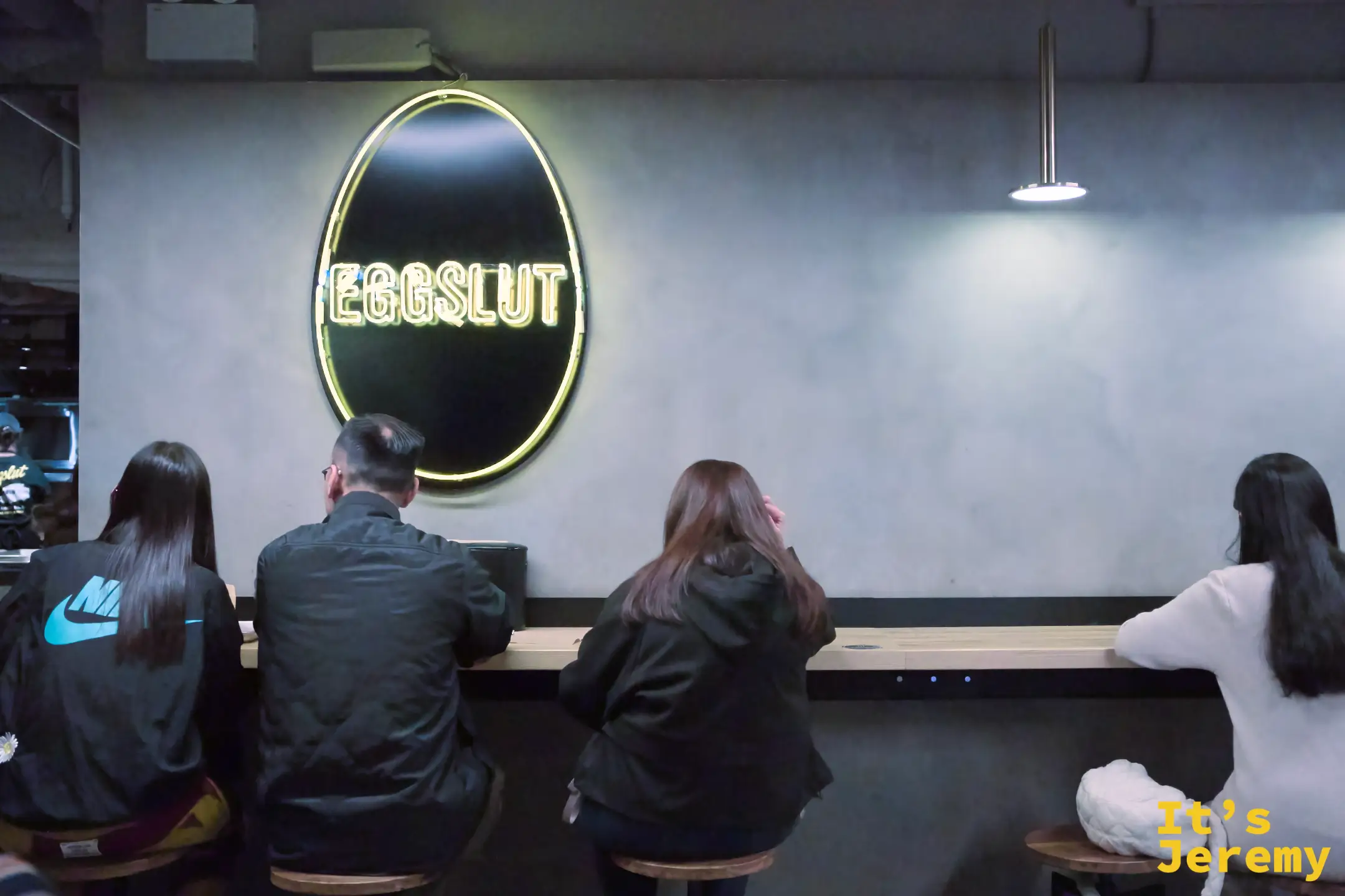 eggslut 店內一面蛋形發光 logo 掛在水泥牆上，下方吧台座位坐著四位客人並肩用餐