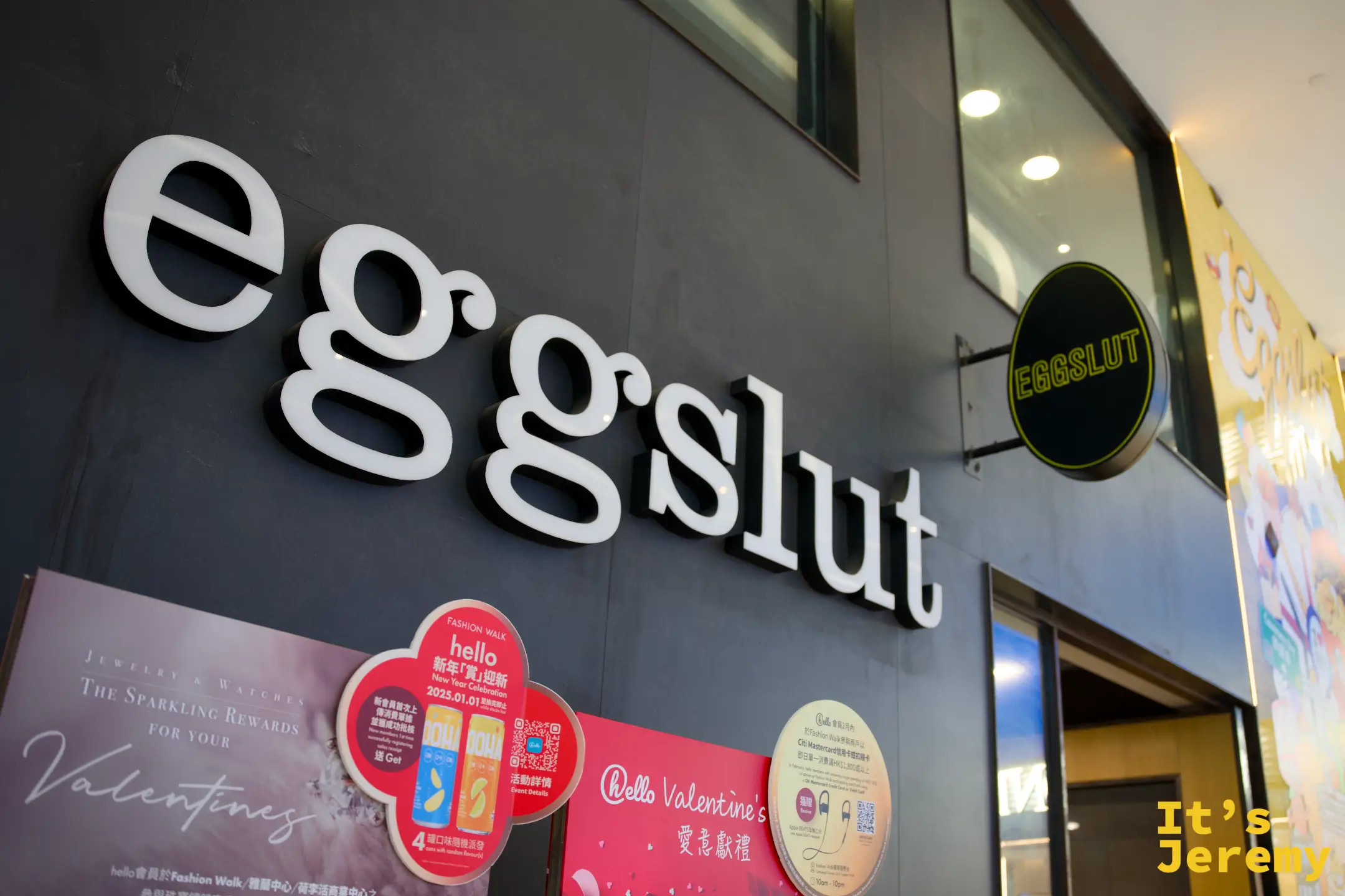 eggslut 商場店面全景，白色「eggslut」招牌旁是一整幅香港主題的彩色壁畫，畫著蛋料理小人、叮叮車與 I♥HK，後方有手扶梯