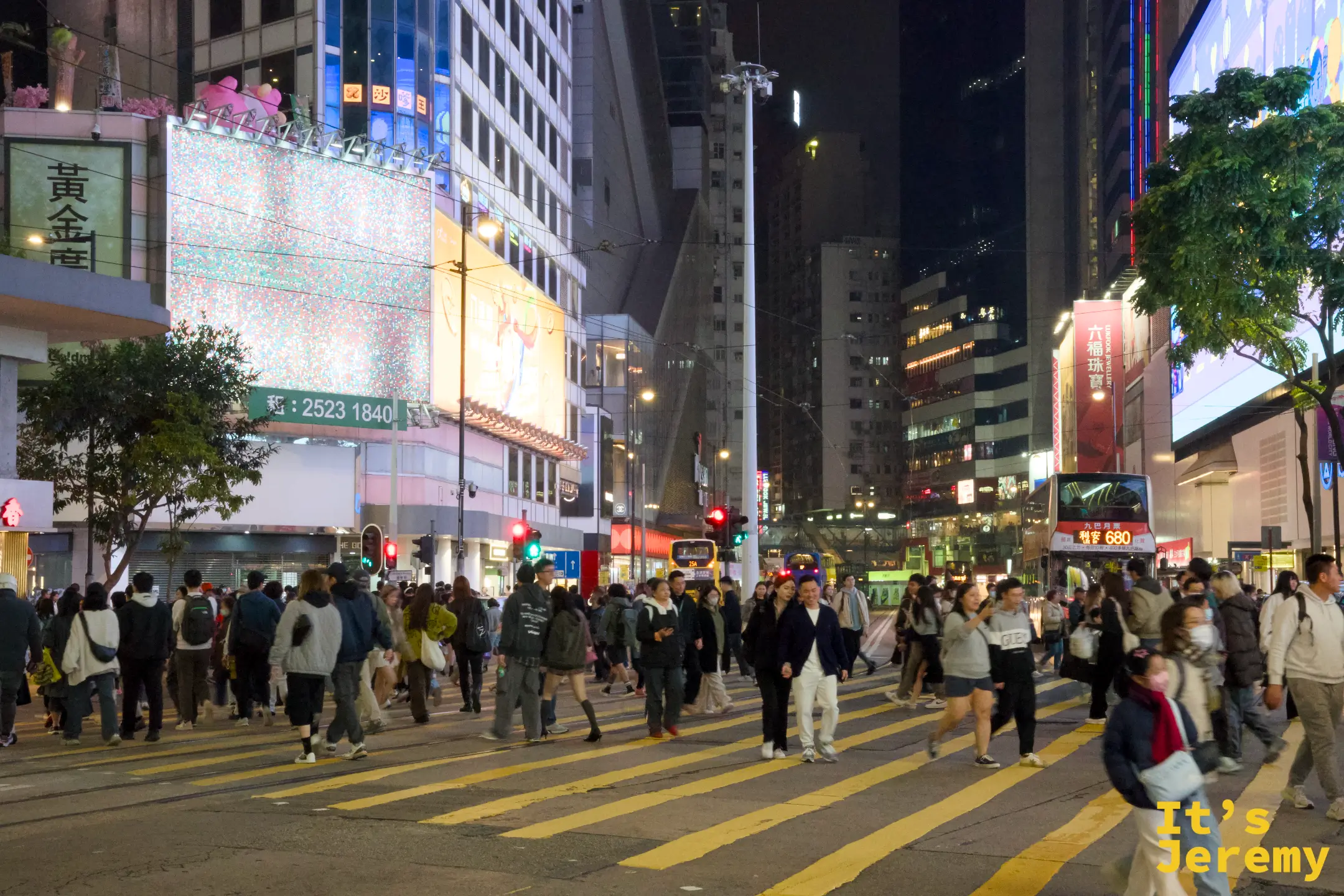 銅鑼灣入夜的十字路口，黃色斑馬線上滿是穿越的人潮，兩旁大樓掛滿霓虹招牌與大型螢幕，遠處有巴士與街燈