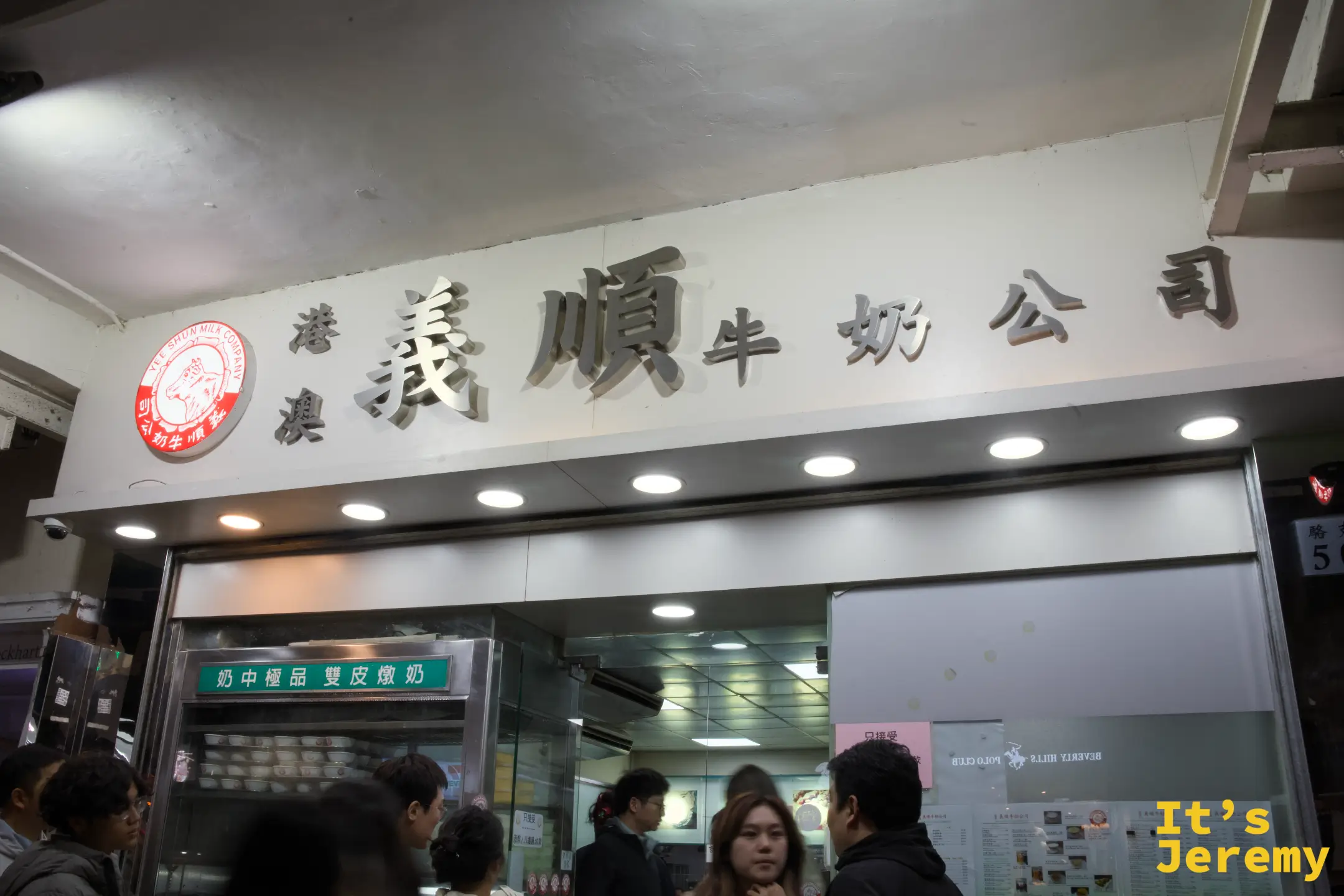 義順牛奶公司店面招牌特寫，白底金屬立體字「港澳義順牛奶公司」與紅色乳牛 logo，玻璃櫃裡排著一碗碗燉奶，門口有客人