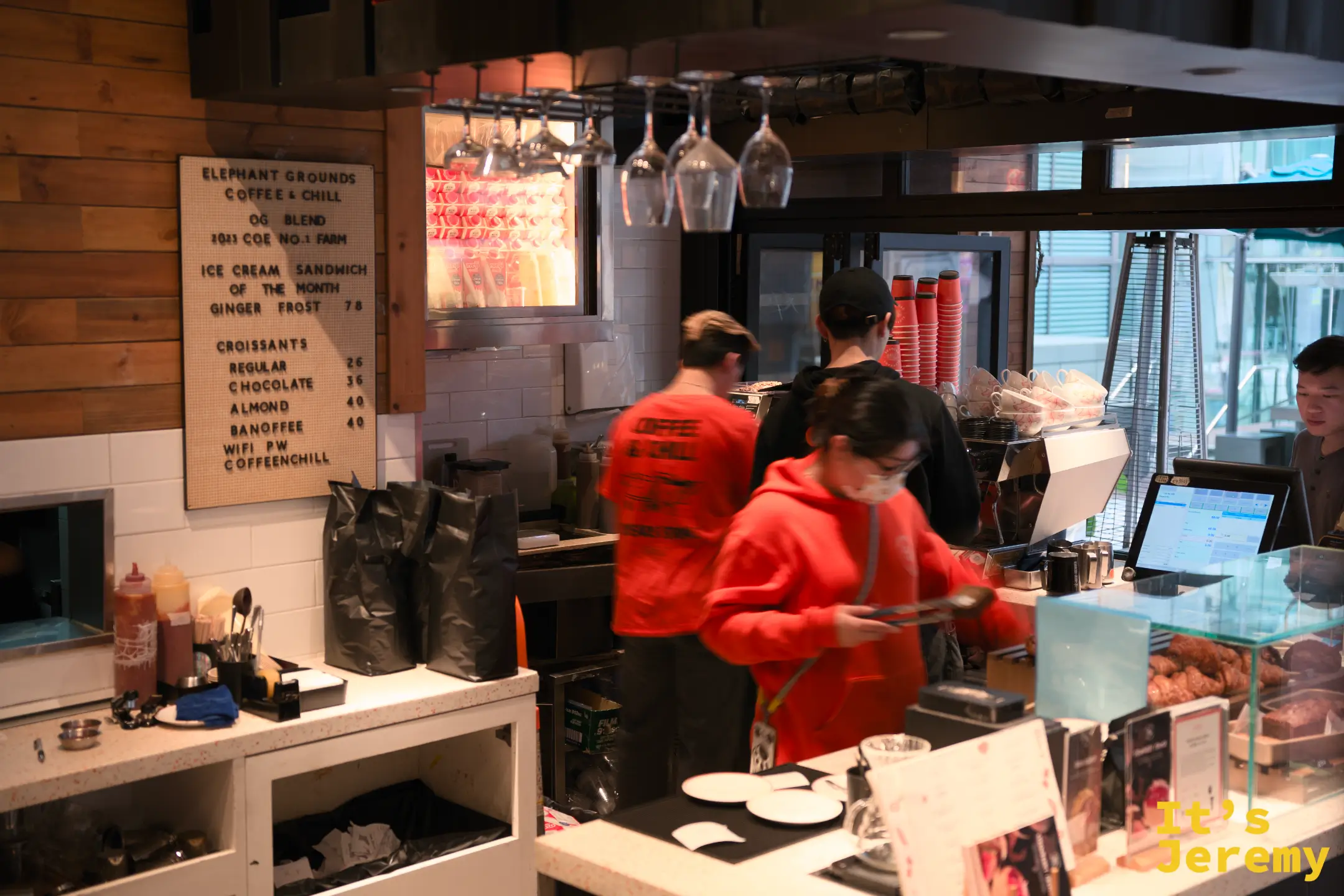 咖啡吧台的廣角，店員與客人交錯，右側是甜點櫃與點餐櫃檯