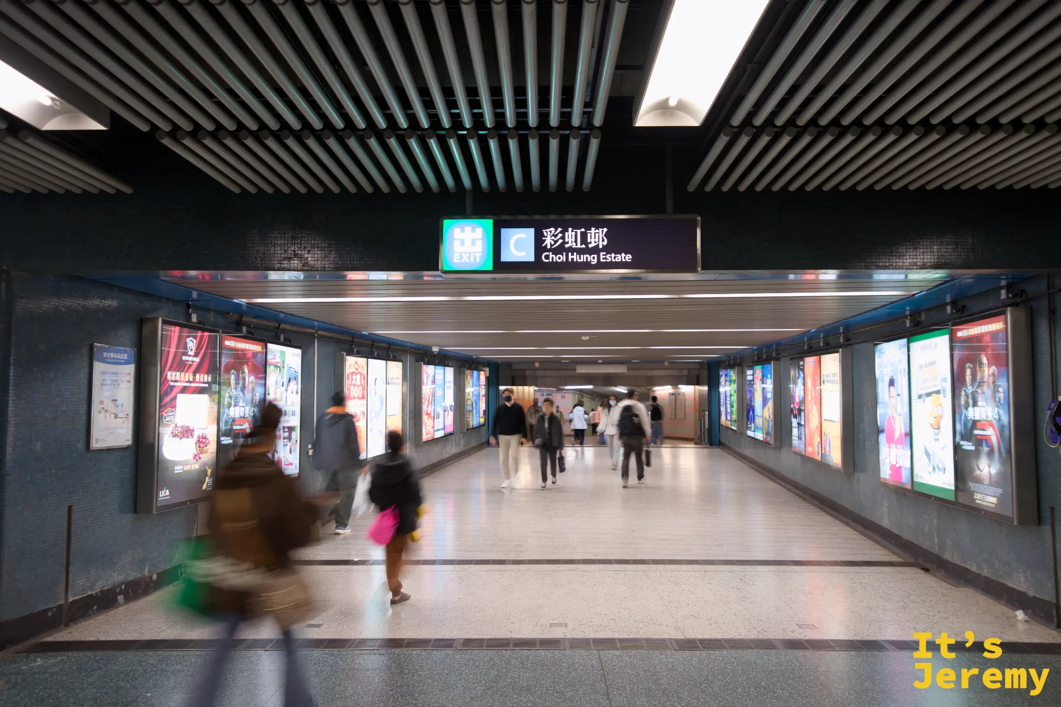 彩虹站的地下通道，天花板懸著「彩虹邨 Choi Hung Estate」的指示牌，兩側是廣告燈箱與往來的人