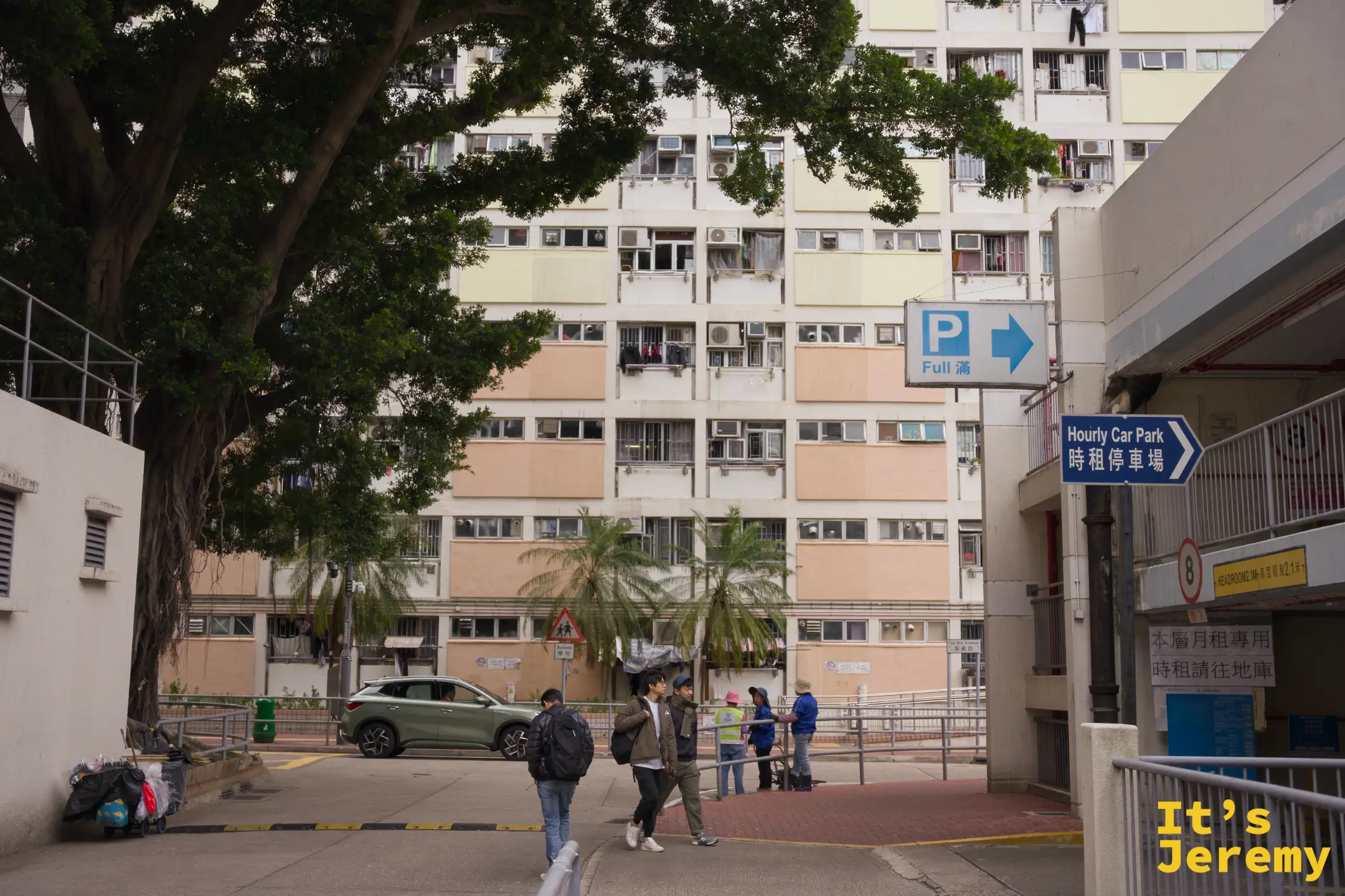 屋邨內的路口，藍色路牌寫著「Hourly Car Park 時租停車場」與「Full 滿」，幾名遊客正走過，背景是粉彩樓群