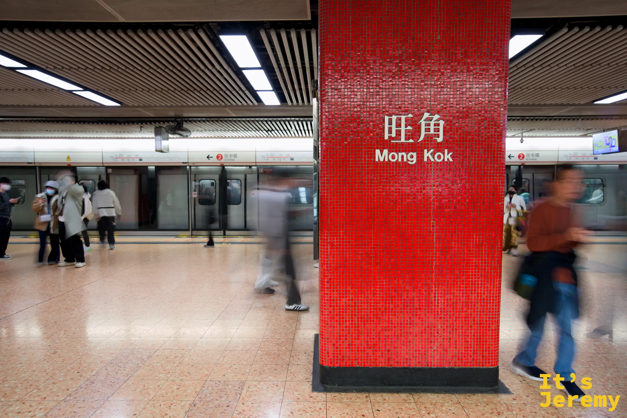 旺角站月台的紅色磁磚立柱，寫著站名「旺角 Mong Kok」，兩側月台有列車與往來乘客