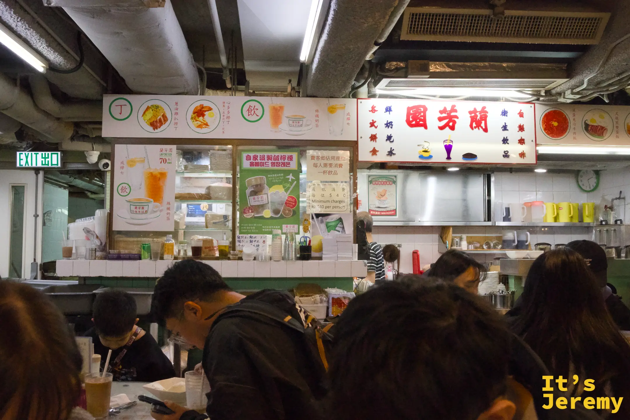 店內開放式的點餐櫃檯，紅色的「蘭芳園」招牌高掛，客人擠滿座位，牆上貼著「每人最低消費一杯飲品」與 EXIT 出口指示