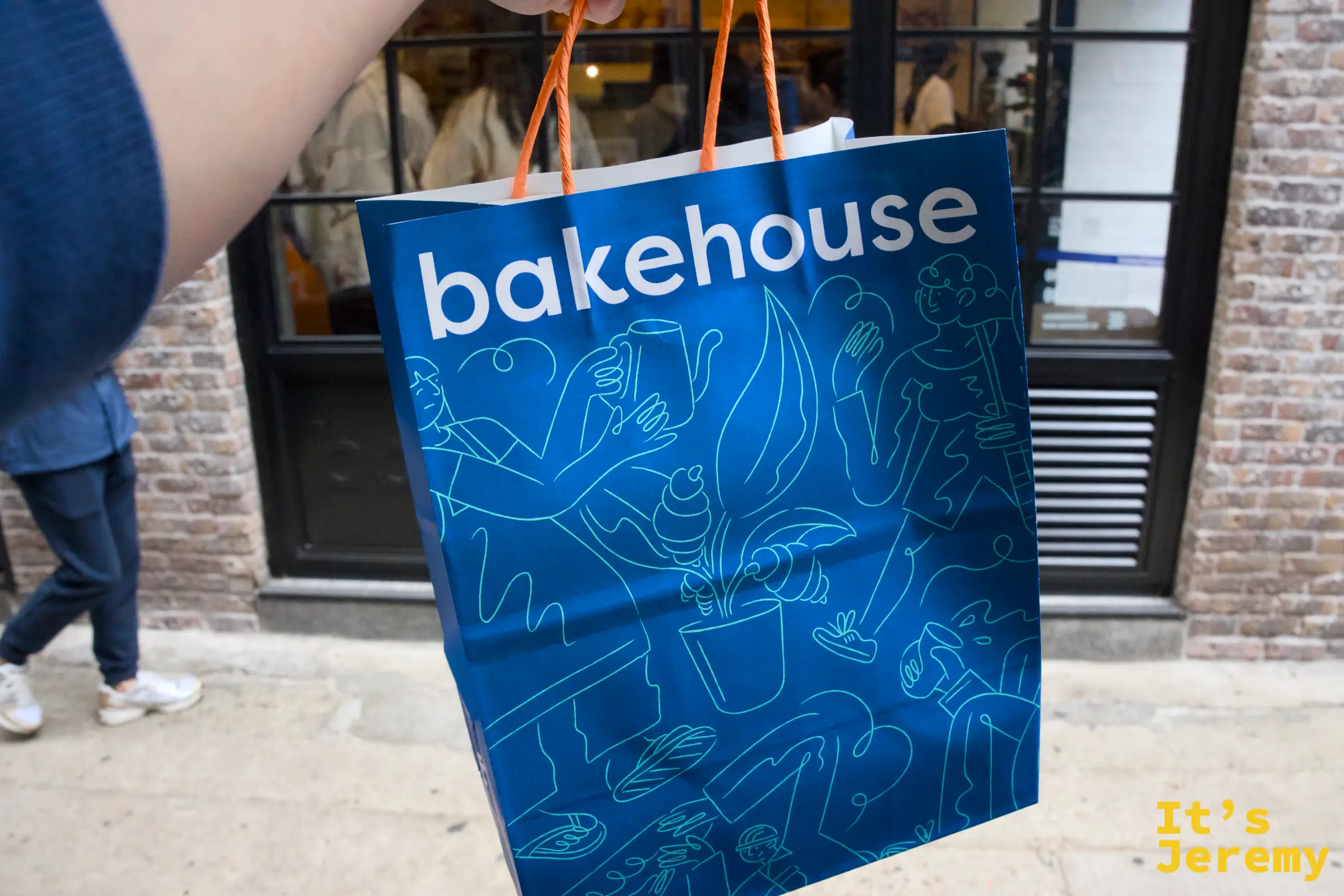 手提著一只印有插畫的藍色「bakehouse」紙袋，背景是店面的磚牆與黑框玻璃窗