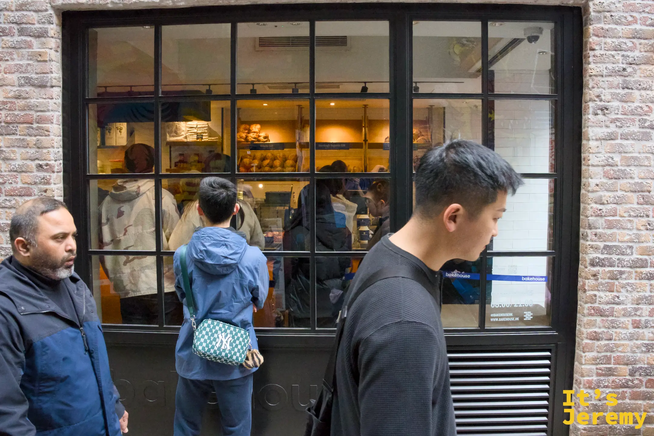 店面的黑框玻璃窗外，幾名客人排隊等候，窗內看得到明亮的烘焙櫃檯與麵包