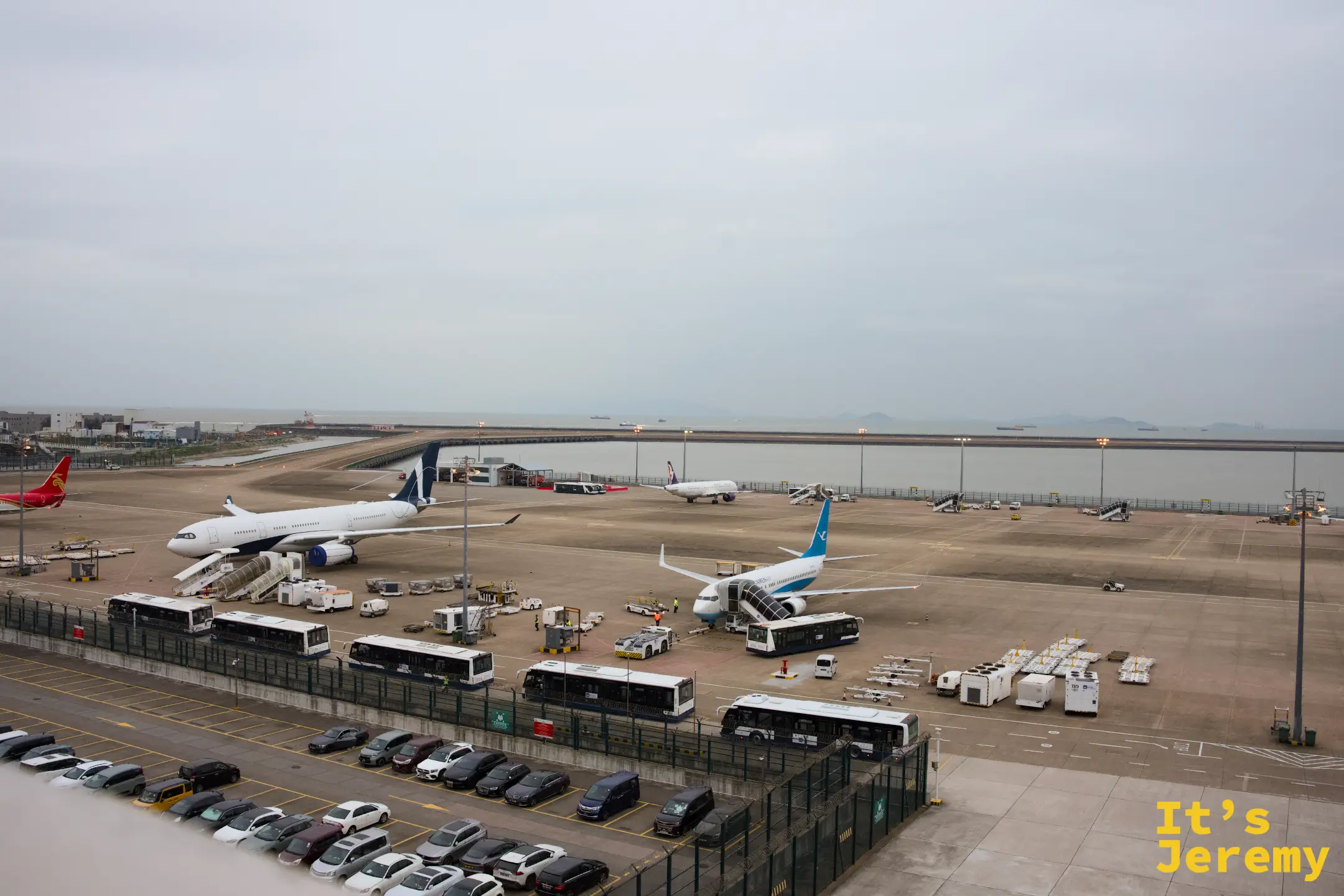 機場停機坪俯瞰，白色客機停靠在空橋與登機梯旁，前方停著一整排接駁巴士，背景是灰白天色下的海與貨船