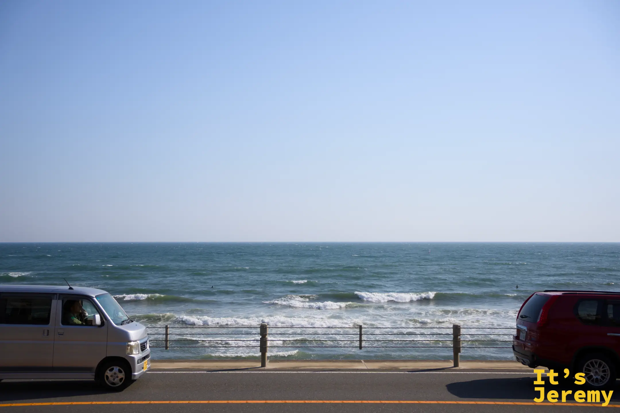 沿著湘南 134 號縣道的海岸線，路上有銀色廂型車與紅色轎車經過，右側就是相模灣