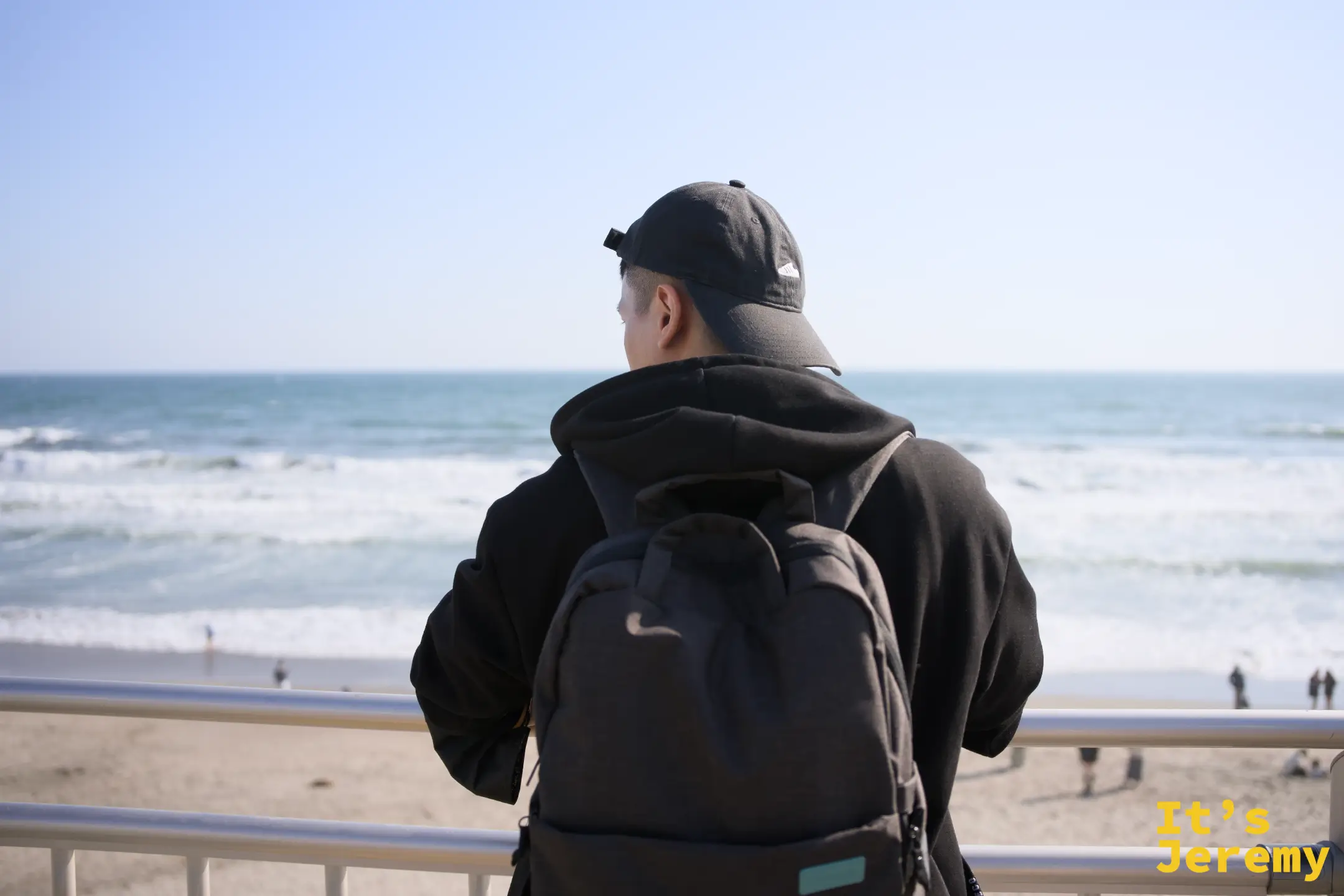 一名背著背包的男子站在海岸欄杆旁，背對鏡頭靜靜望著相模灣