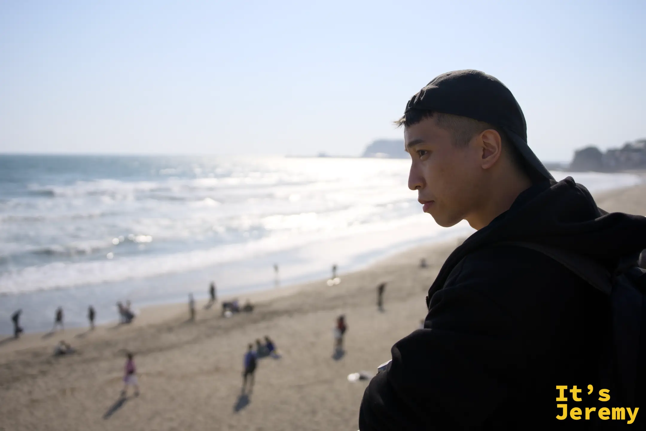 一名戴黑色帽子的男子側臉望向遠方，背後是沙灘與零星散步的人影