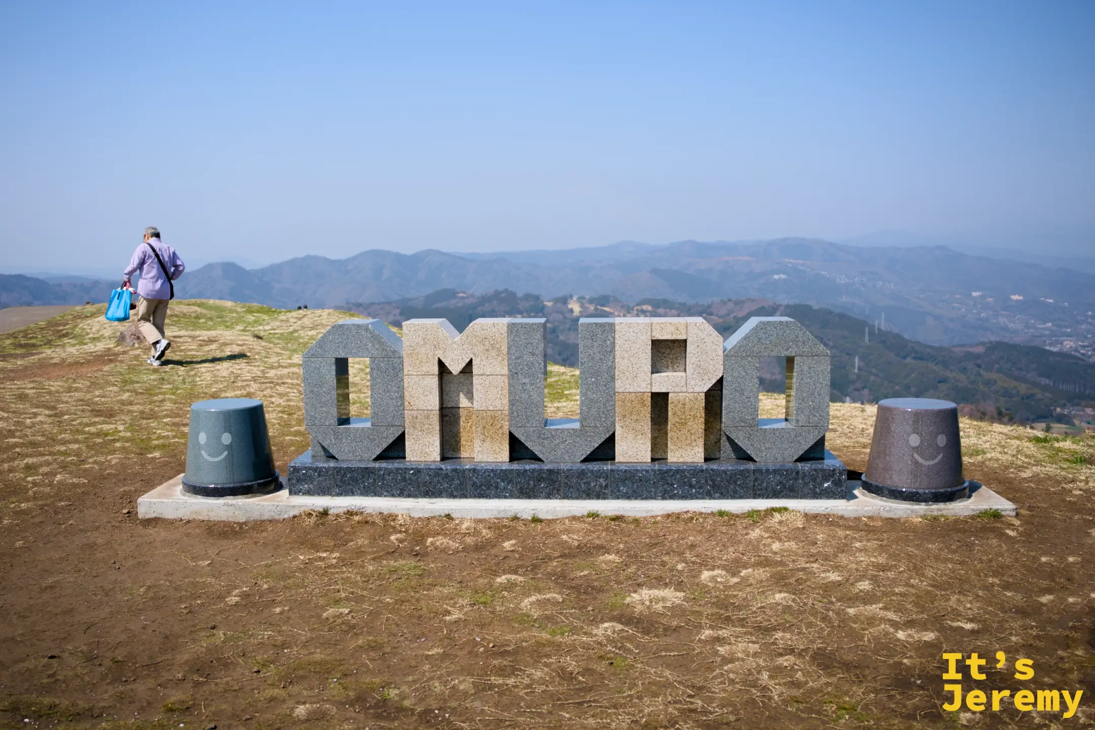 山頂 OMURO 石刻字裝置，兩側有笑臉小石柱，背景為伊豆山脈遠景