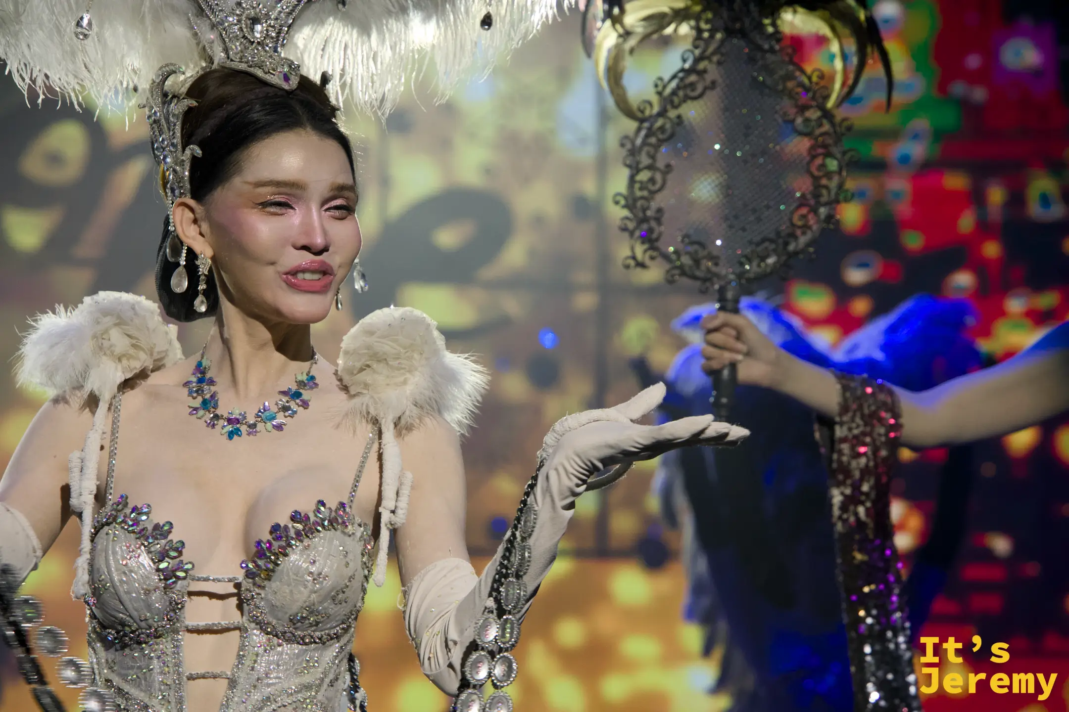金東尼人妖秀舞台上，舞者身穿華麗的銀白色亮片羽毛裝，神情生動地向台下觀眾招手互動。