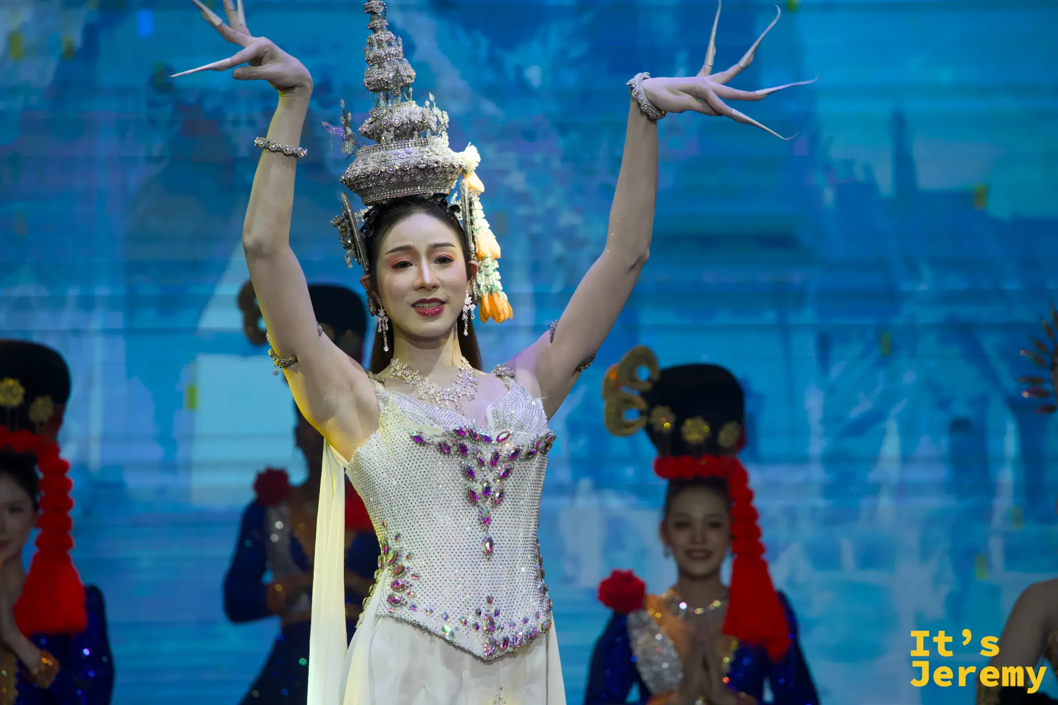 金東尼人妖秀舞台上，舞者身穿純白亮片禮服、頭戴泰式傳統尖頂冠冕，雙手擺出優雅的古典舞蹈手勢。