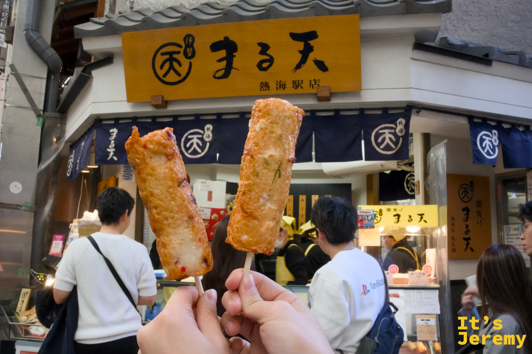 兩根炸竹輪棒舉高在まる天熱海店招牌前，橘色暖簾與店名清晰可見
