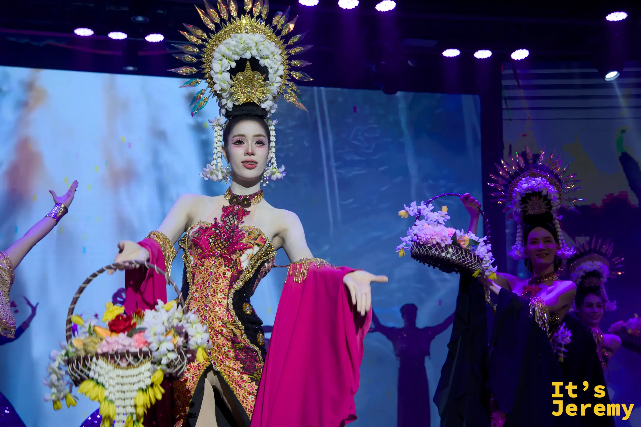 金東尼人妖秀舞台上，舞者身穿桃紅與金色相間的華麗禮服，頭戴誇張的圓形放射狀花藝頭飾，手挽花籃進行走秀。