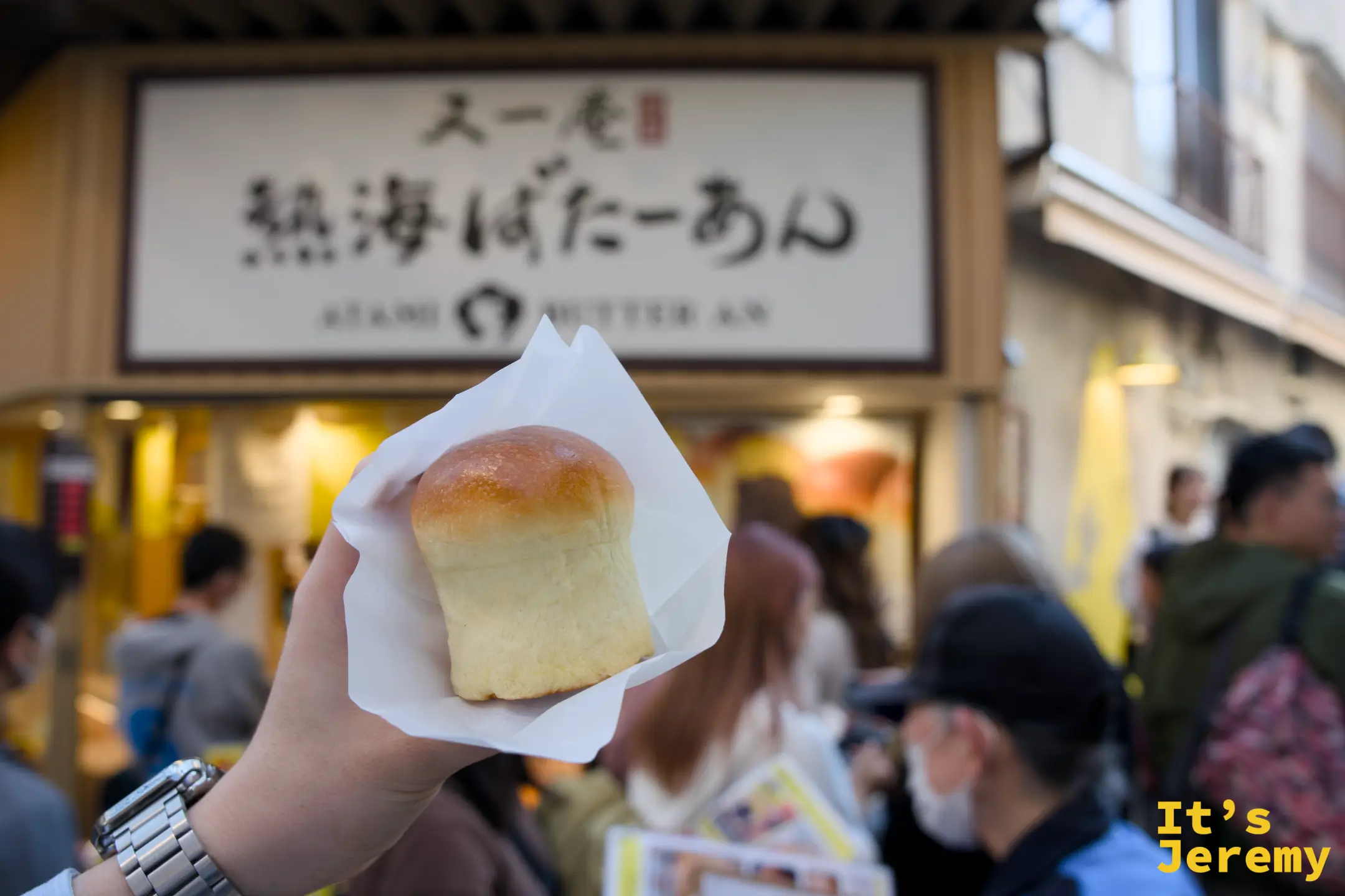 手持奶油饅頭高舉在又一庵店頭前，背景為熱海ばたーあん招牌與店門口排隊人潮