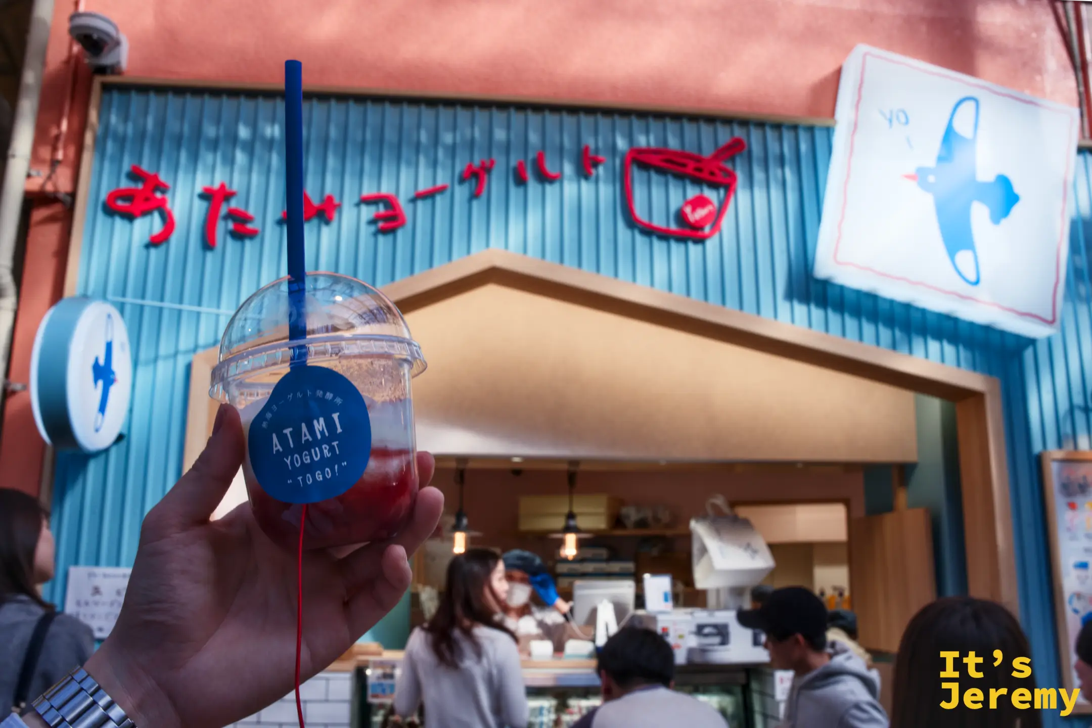 手持 ATAMI YOGURT 藍色貼紙飲品杯，背景為粉橘系外牆與紅字あたみヨーグルト招牌