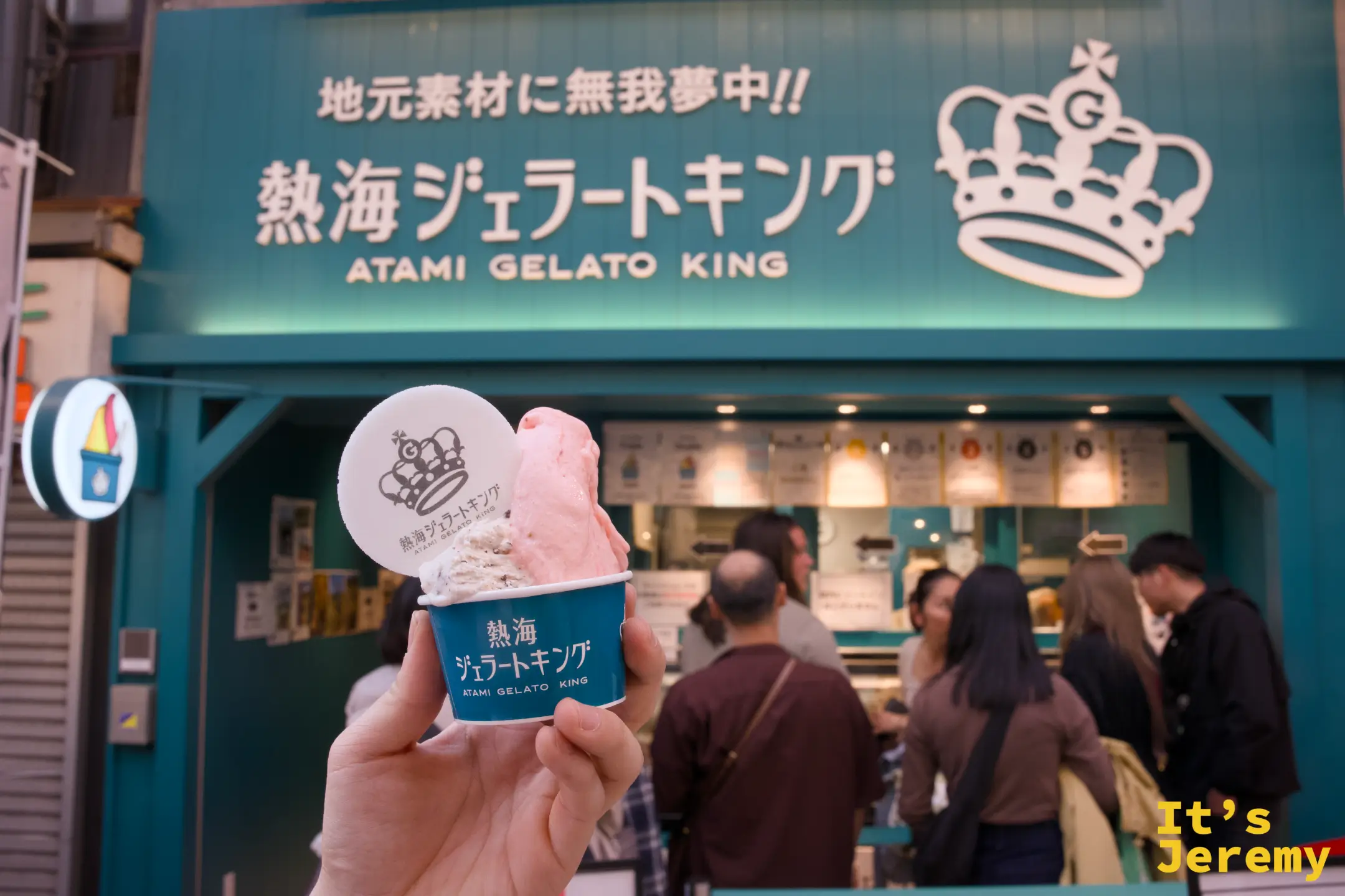 手持熱海ジェラートキング雙球冰淇淋杯，背後為綠色店面招牌與皇冠 LOGO，店內仍有多人排隊