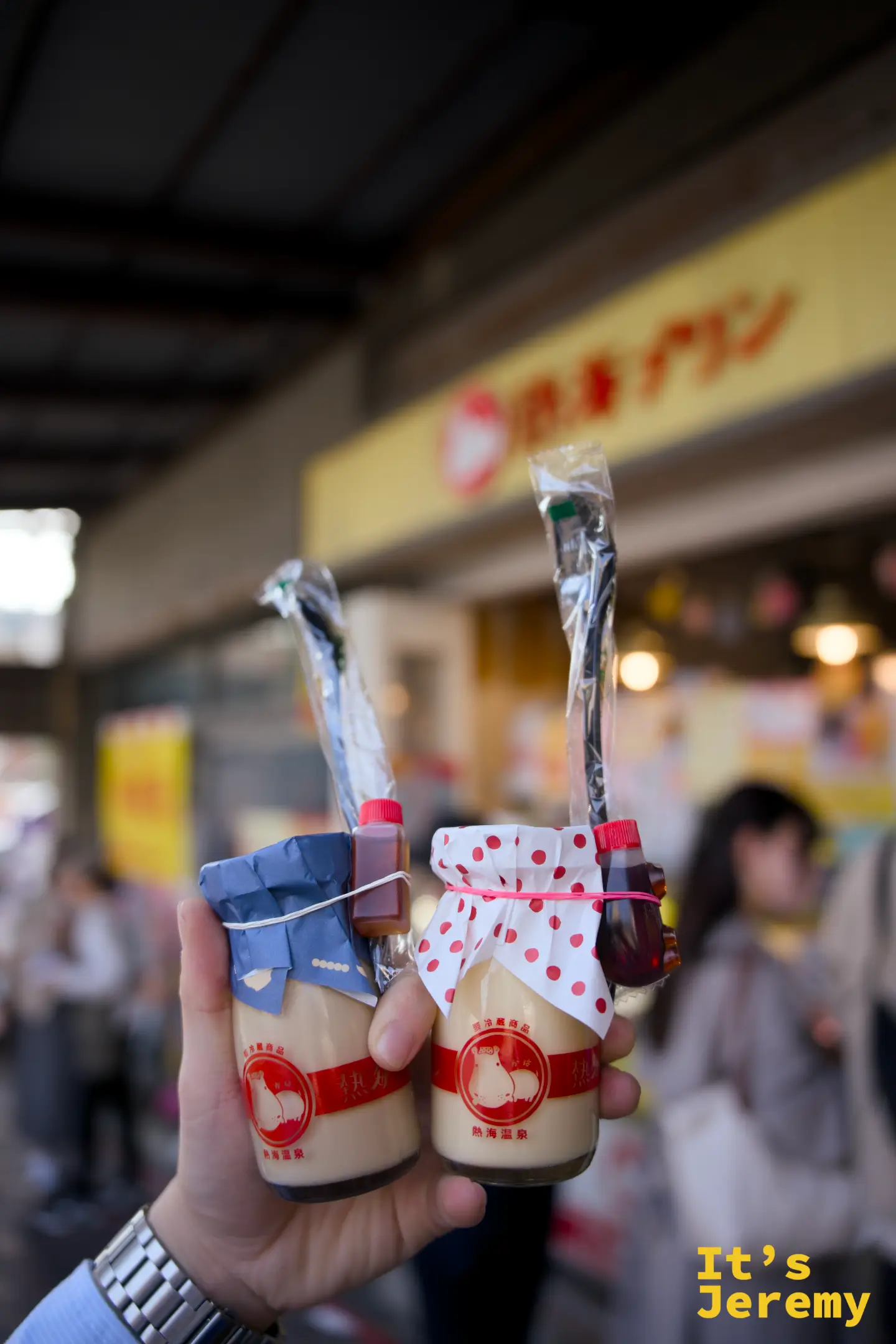 雙手各持一瓶熱海プリン，左為藍色、右為白色圓點包裝，各附一小瓶焦糖醬