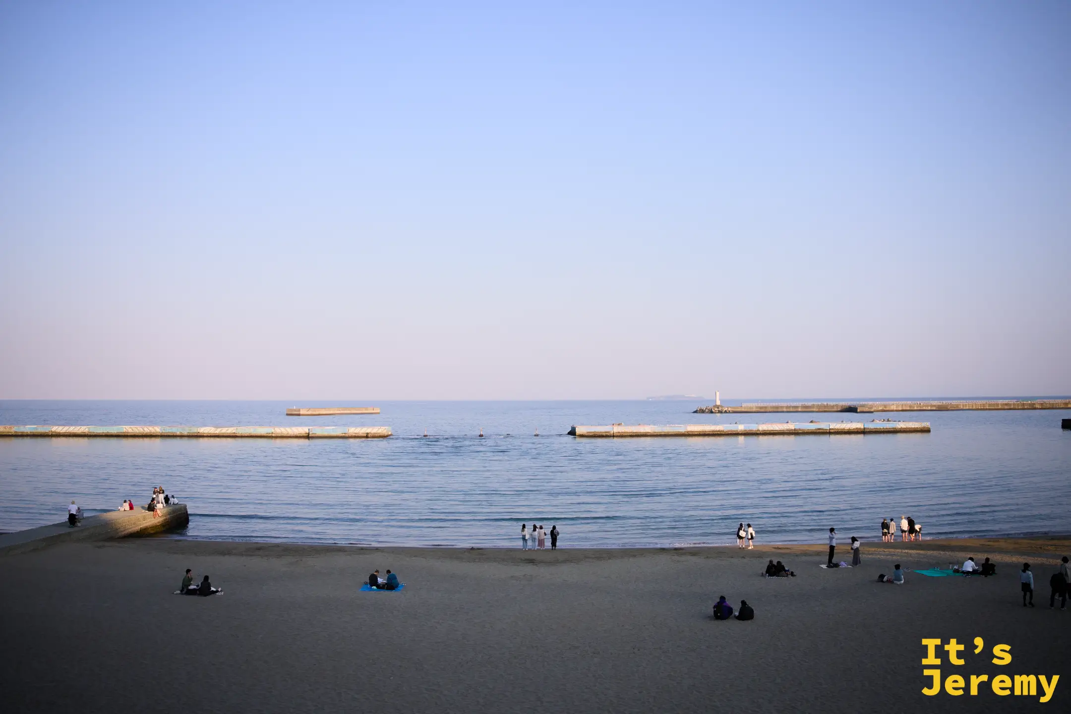 熱海砂浜全景，天色泛藍粉暮色，遠方防波堤與燈塔清晰，沙灘上三三兩兩的人群席地而坐