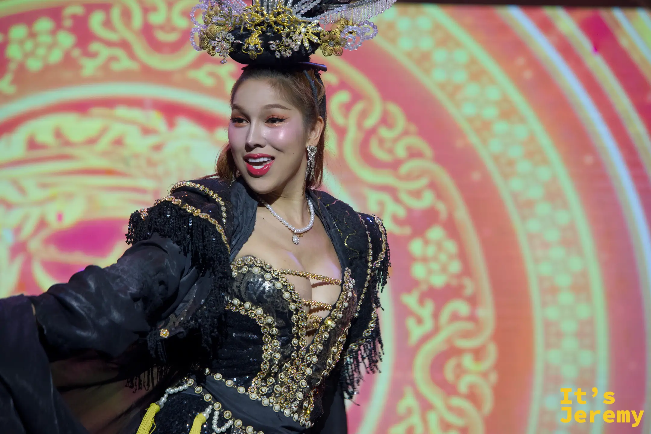 金東尼人妖秀舞台上，舞者身穿綴有金色亮片的黑色中式風馬甲禮服，搭配精緻髮冠，在喜慶的紅金背景前熱情舞動。